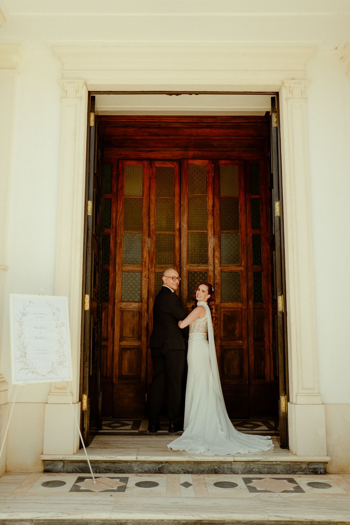 Bride and groom standing in elegant wooden doorway of wedding venue, bride in white gown with train.
