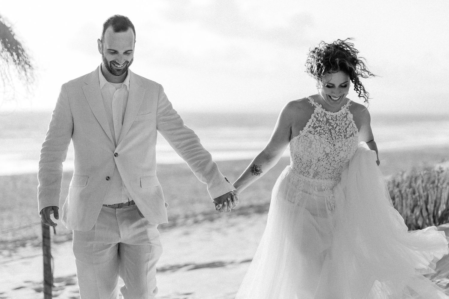 Black and white photo of a bride and groom holding hands and walking on a beach in wedding attire.