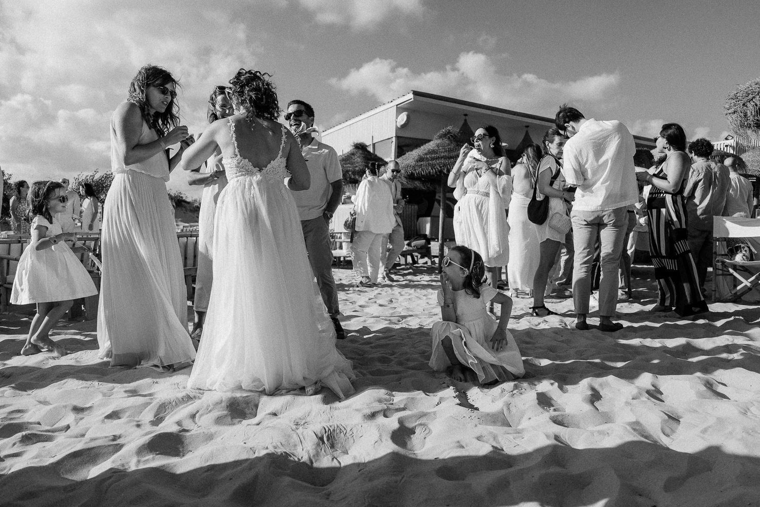 Black and white beach wedding scene with bride, guests, and children gathered on sandy shore near a decorated outdoor venue.