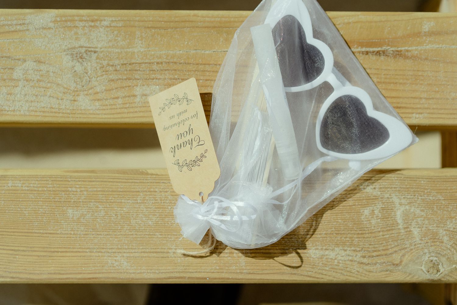 Heart-shaped wedding favor in white tulle bag with kraft paper tag on wooden bench.