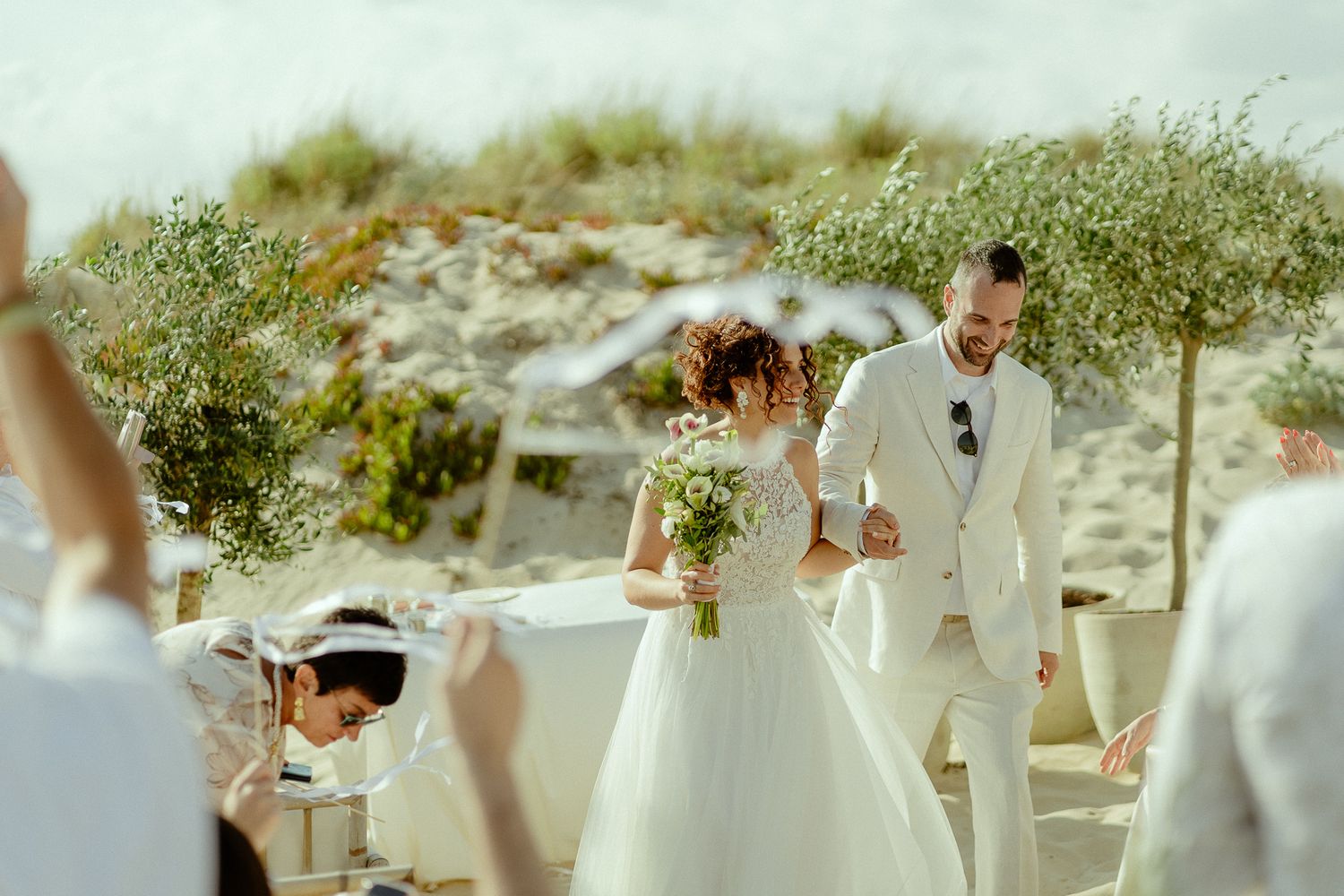 Bride and groom smiling at outdoor beach wedding ceremony with olive trees and guests in background.