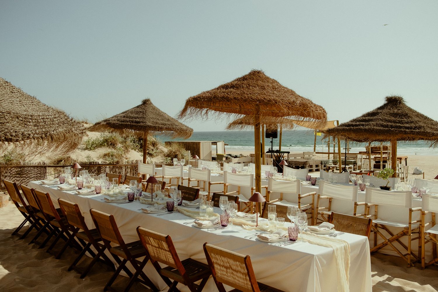 Elegant beachside dining setup with long white tables, thatched umbrellas, and ocean views for a wedding reception.