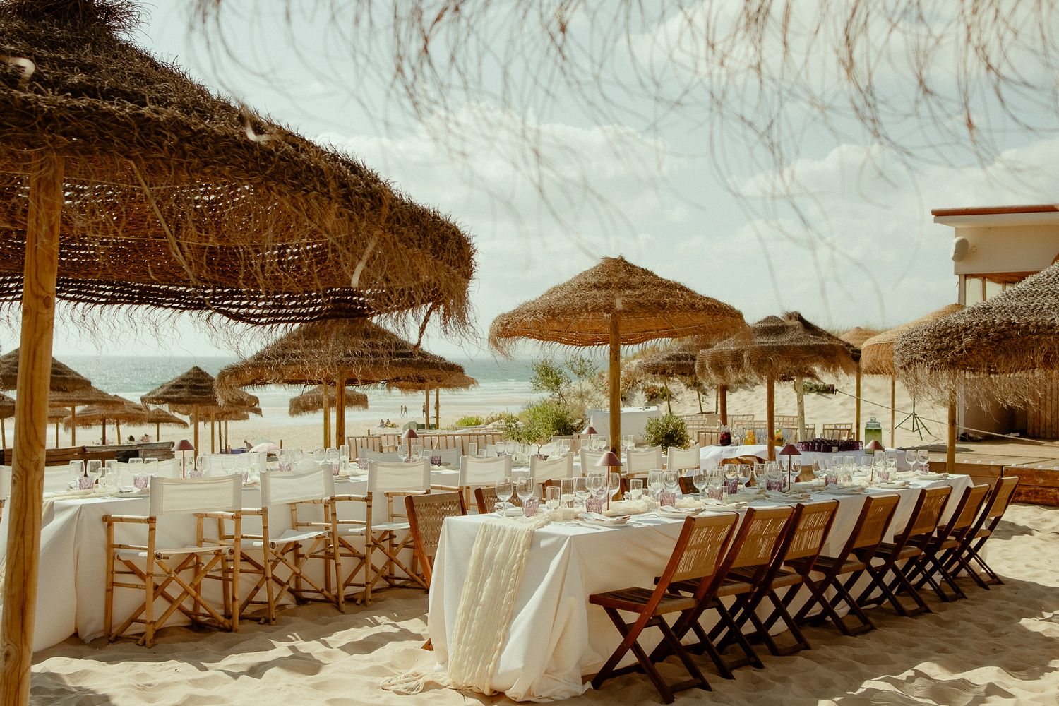 Elegant beach wedding reception setup with thatched umbrellas, white draped tables, and bamboo chairs on sandy shore.