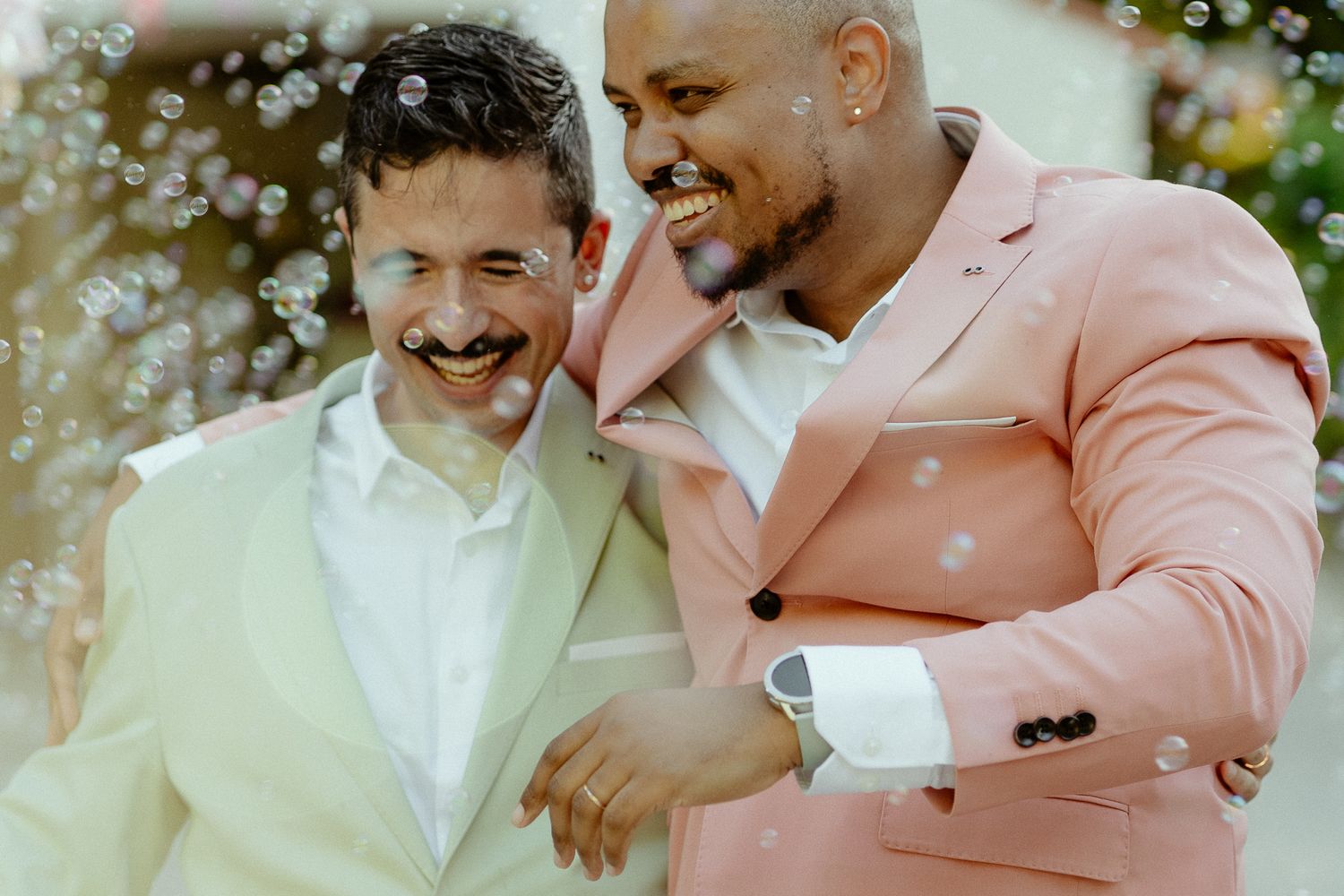 Two grooms laughing joyfully at their wedding, one in white suit and one in pink suit, surrounded by bubbles.