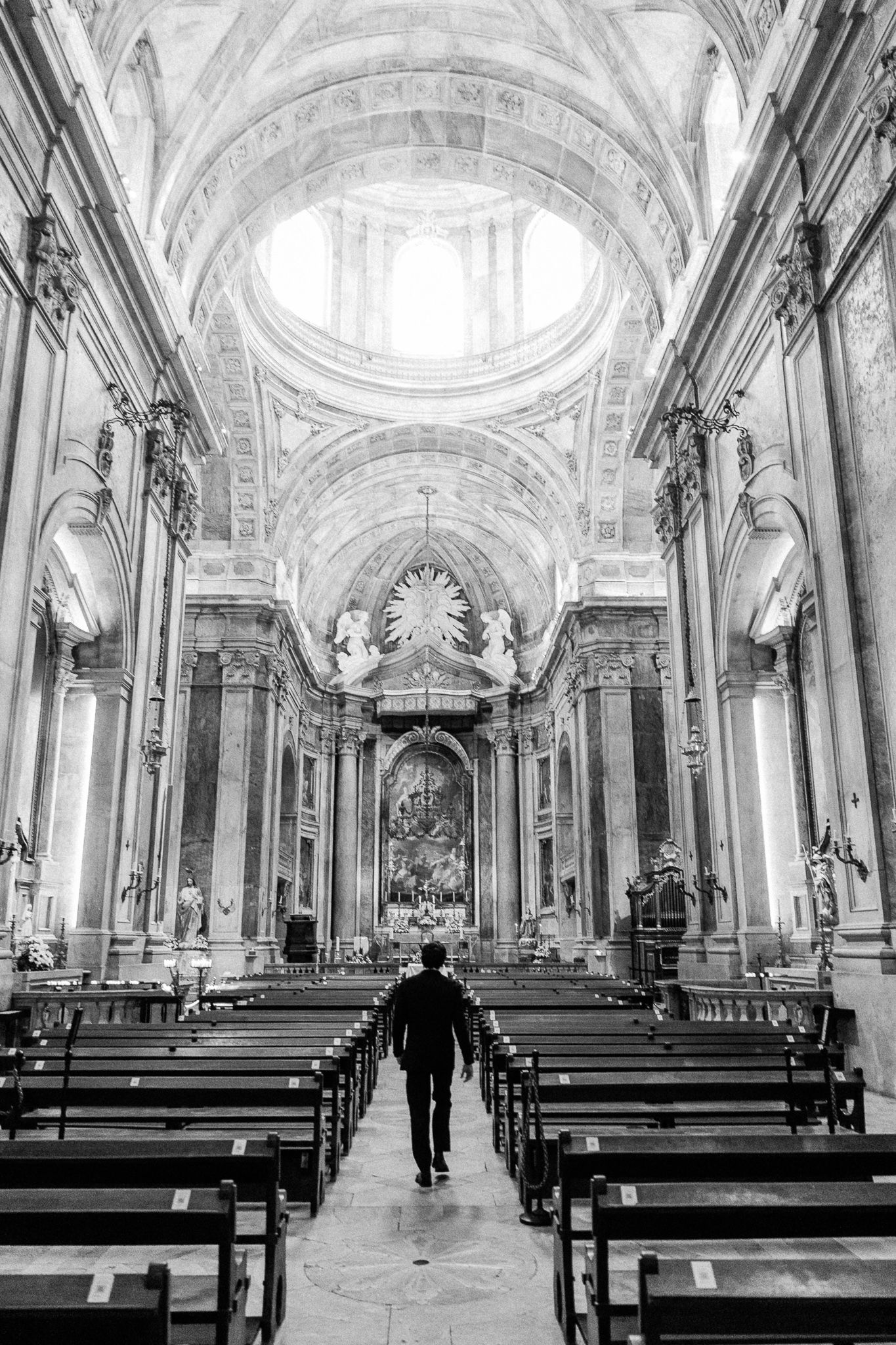 Black and white photo of a man walking down the aisle of an ornate baroque cathedral with grand arches.