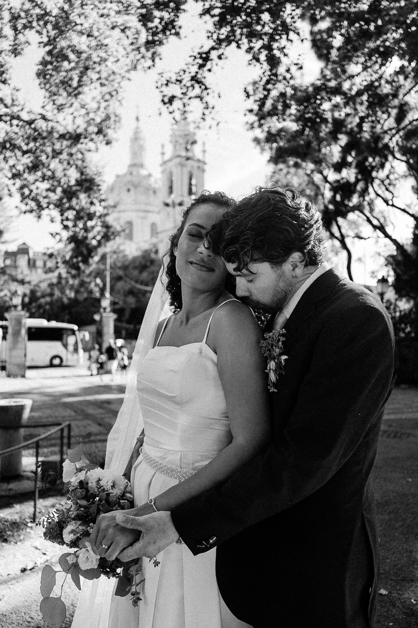 Black and white wedding photo of bride and groom embracing outdoors, with a historic church and trees in the background.