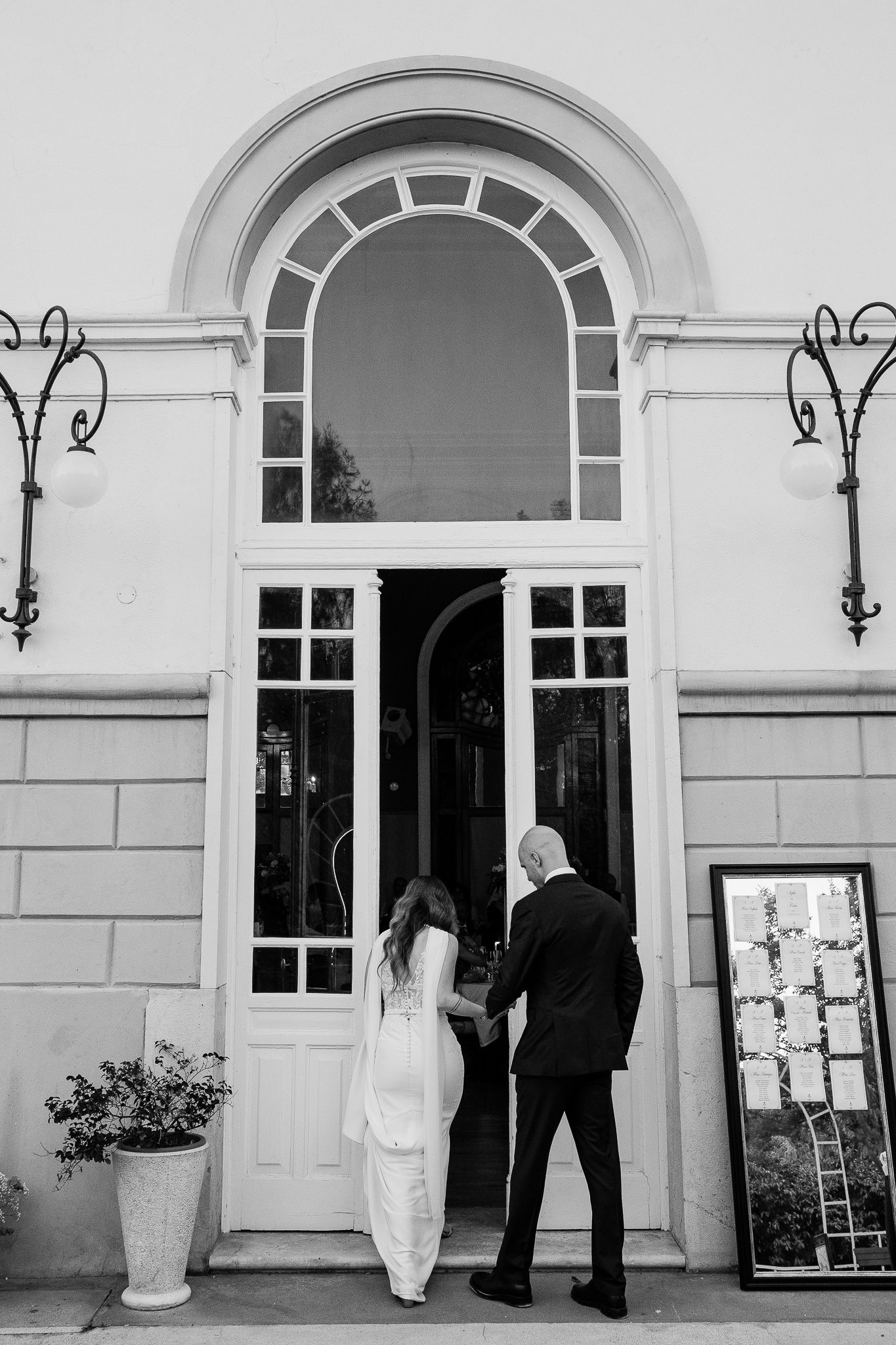 Black and white photo of a bride and groom holding hands entering an elegant venue with arched windows.