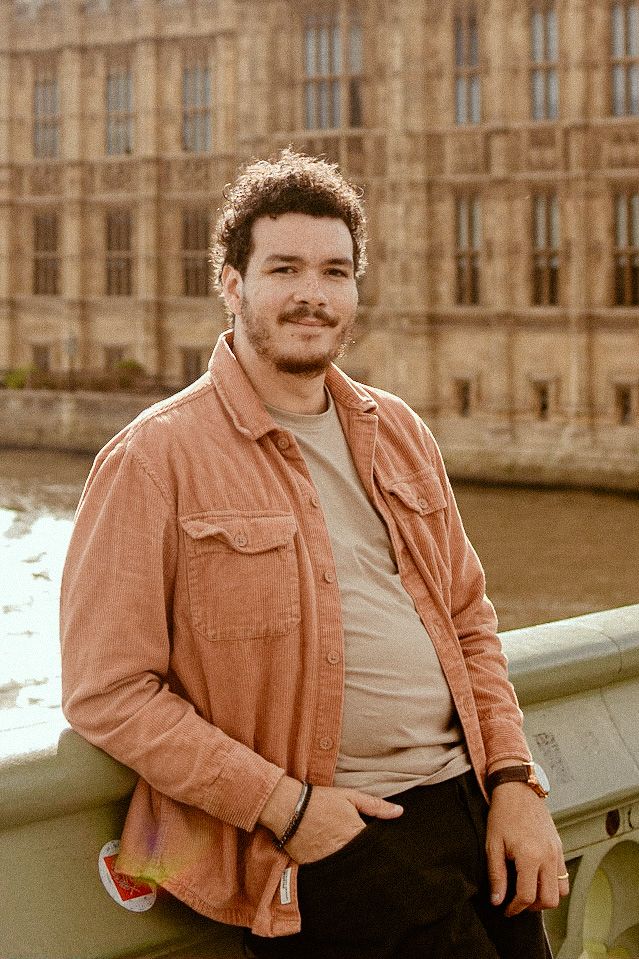 Person poses on bridge railing near historic Parliament building in warm sunset lighting wearing brown jacket.