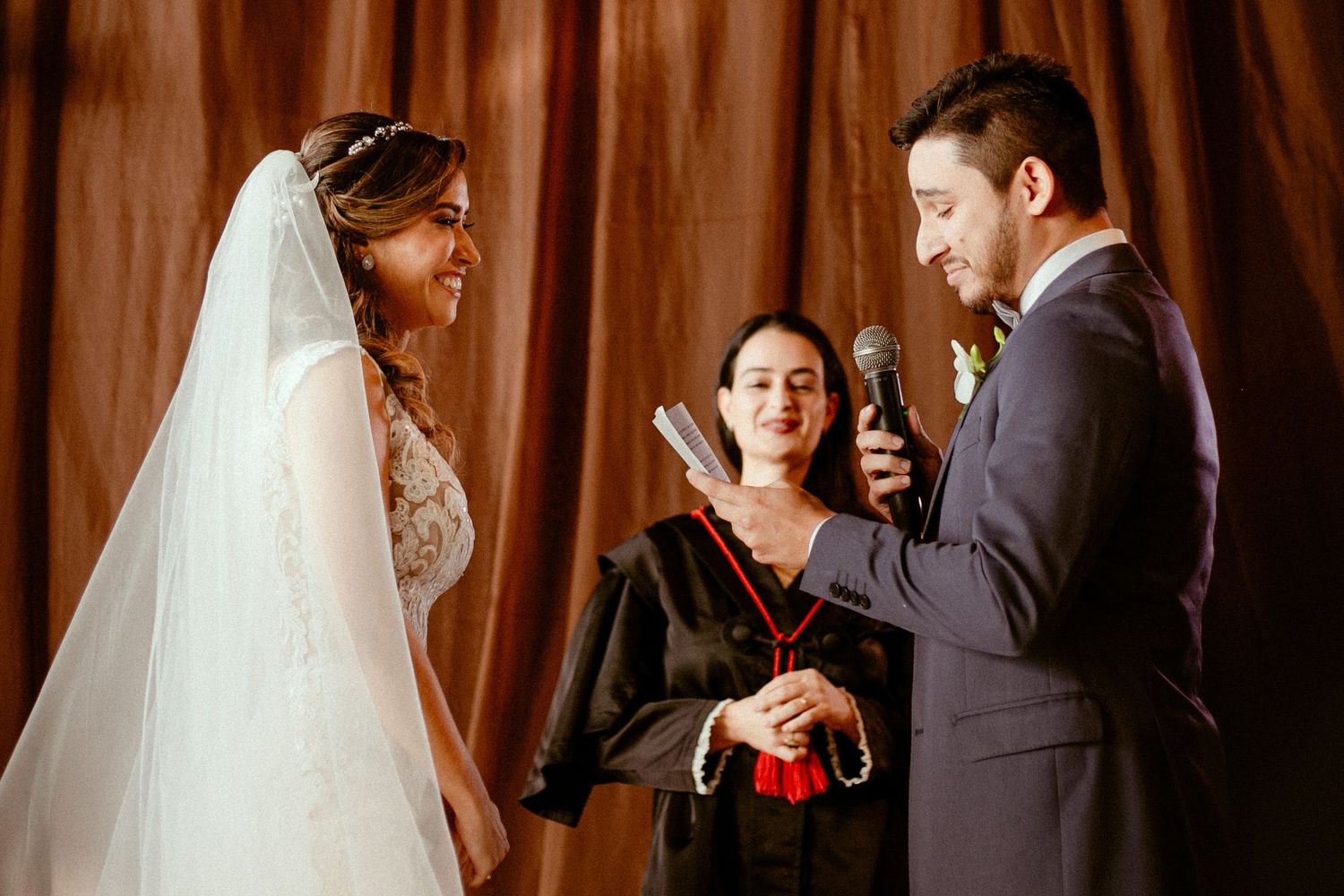 Bride and groom exchange wedding rings during intimate ceremony in front of wooden backdrop.