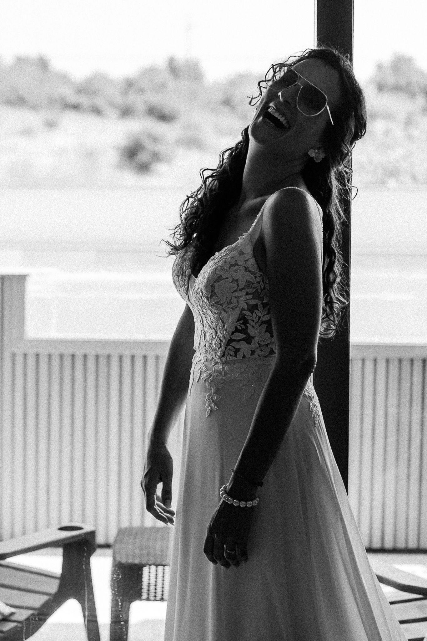 Black and white photo of a laughing bride in a lace wedding gown and sunglasses, posing on a porch.