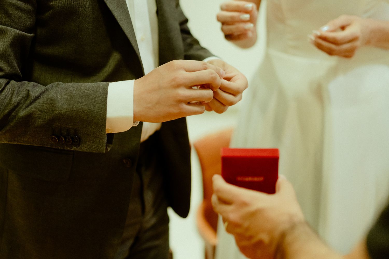 Groom placing a wedding ring on bride's finger during ceremony, with red ring box held by attendant.