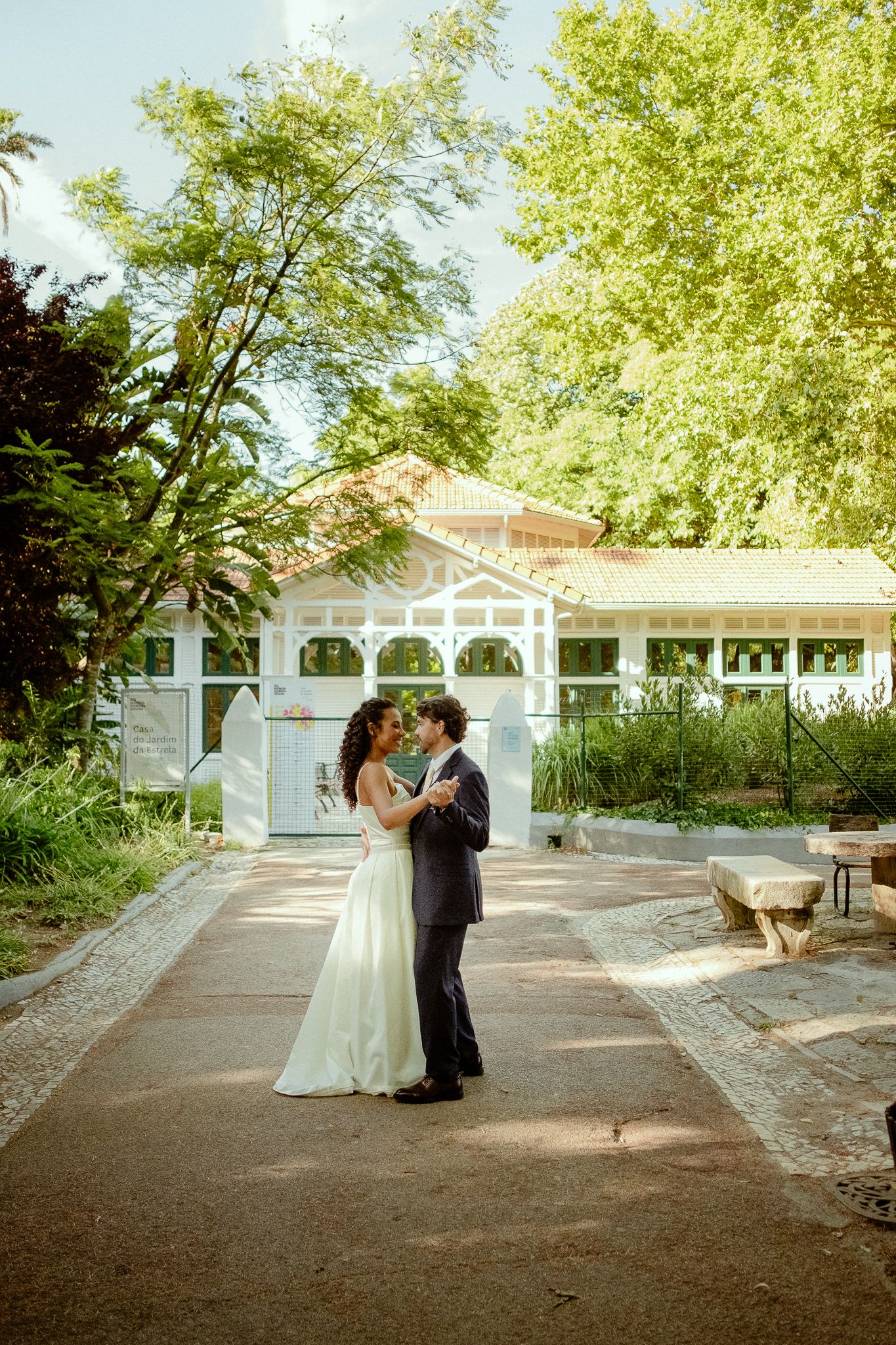 A bride and groom share a romantic embrace in a sunlit garden with a white gazebo and lush green trees.