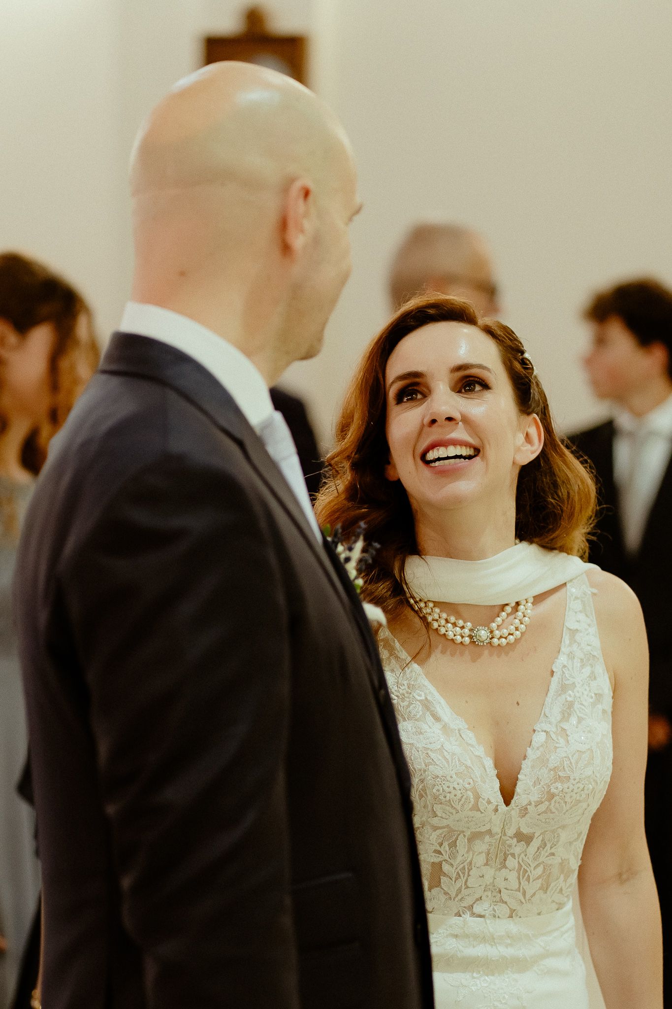 Bride in lace gown and pearl necklace smiling joyfully at groom in black suit during wedding ceremony.