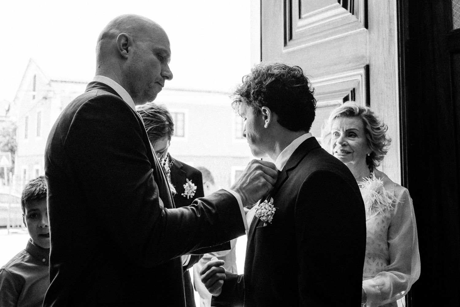Black and white wedding photo of groom having boutonniere pinned while bride watches joyfully in elegant lace gown.