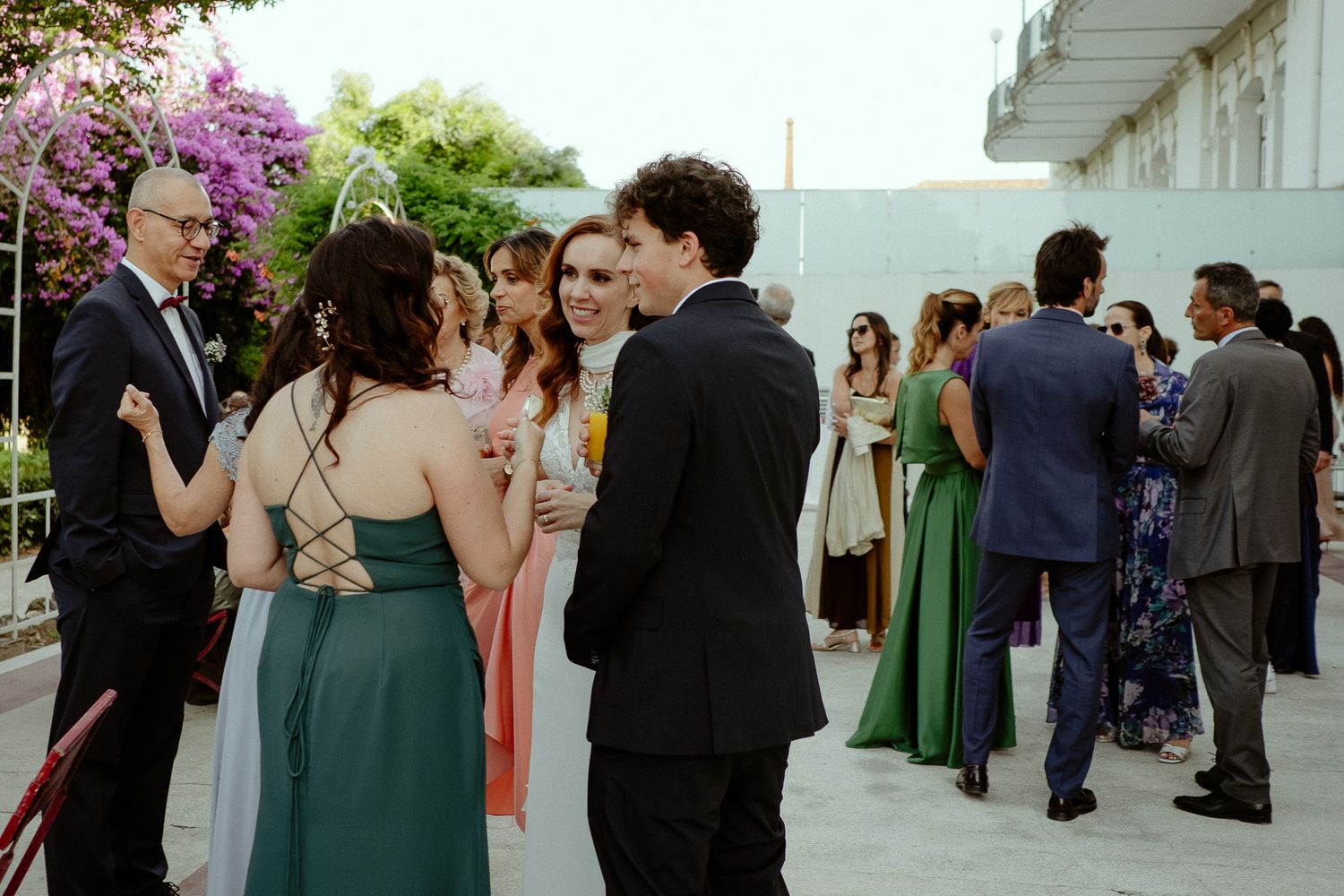 Elegantly dressed wedding guests mingle outdoors, wearing formal attire in green, black, and pastel tones.