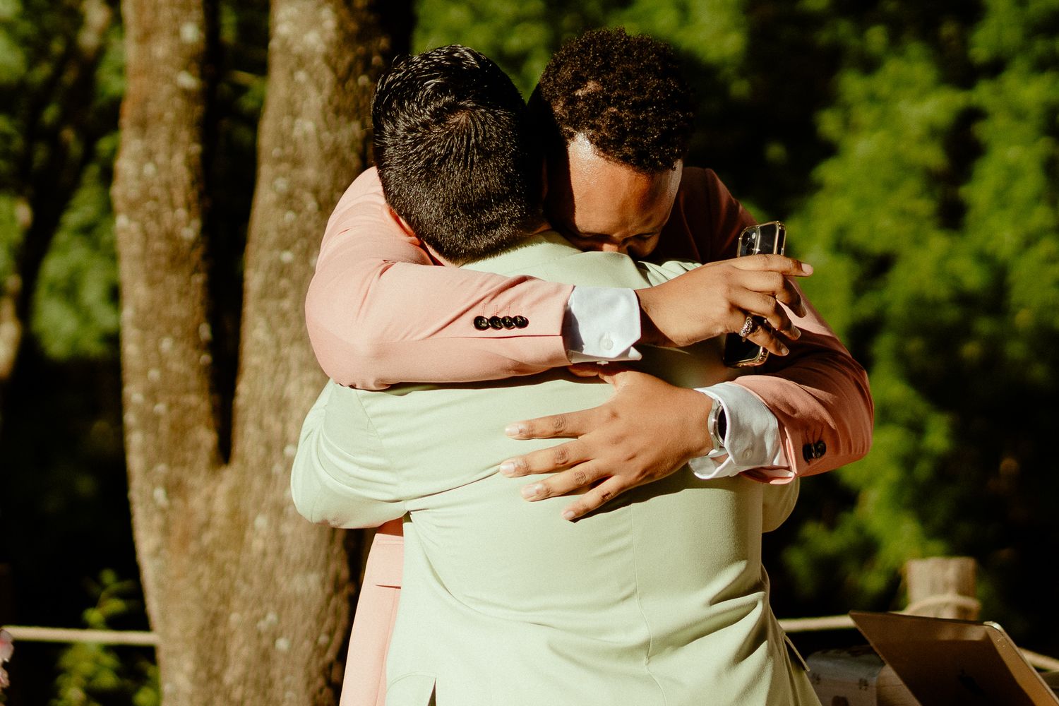 Two men in suits share a heartfelt hug outdoors at a wedding celebration surrounded by lush green trees.