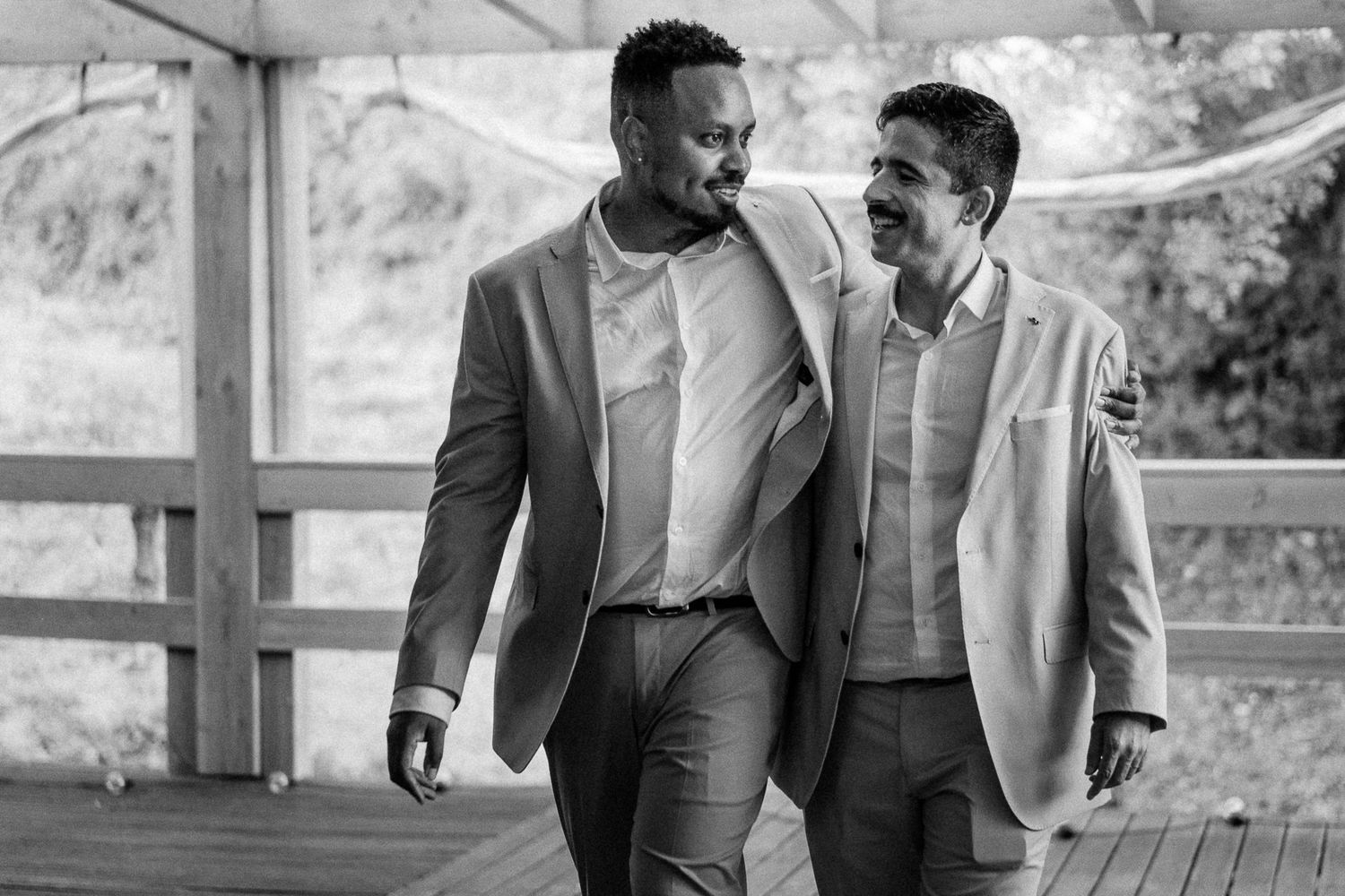 Two men in suits walking together arm in arm, smiling and laughing in a black and white outdoor photo.