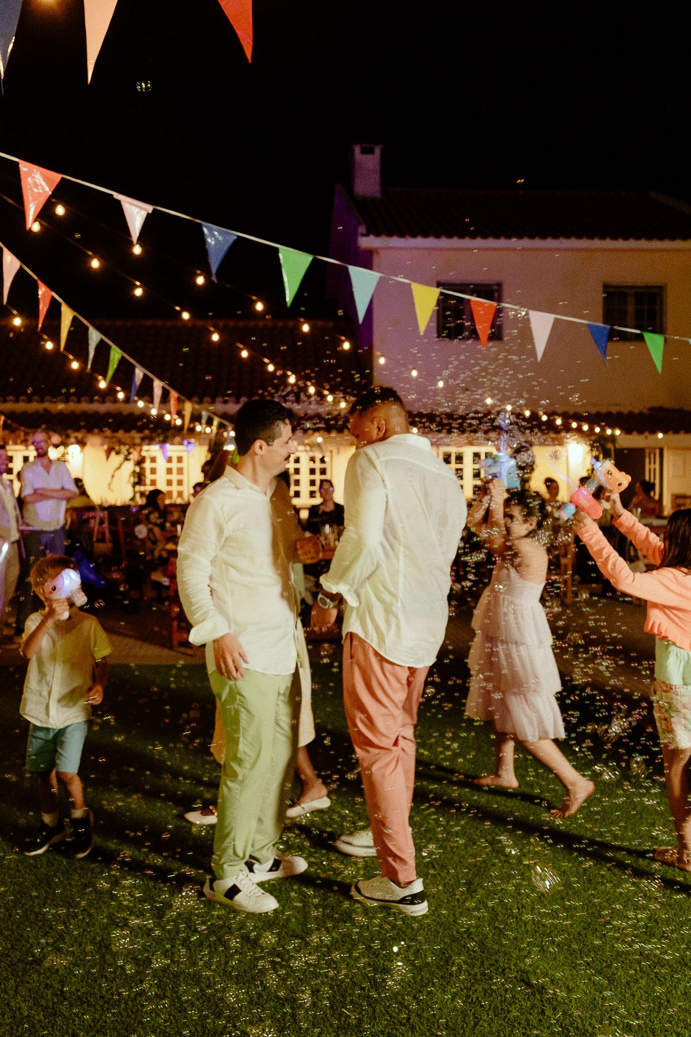 Two grooms in white shirts dance outdoors at night wedding reception with colorful bunting and string lights.