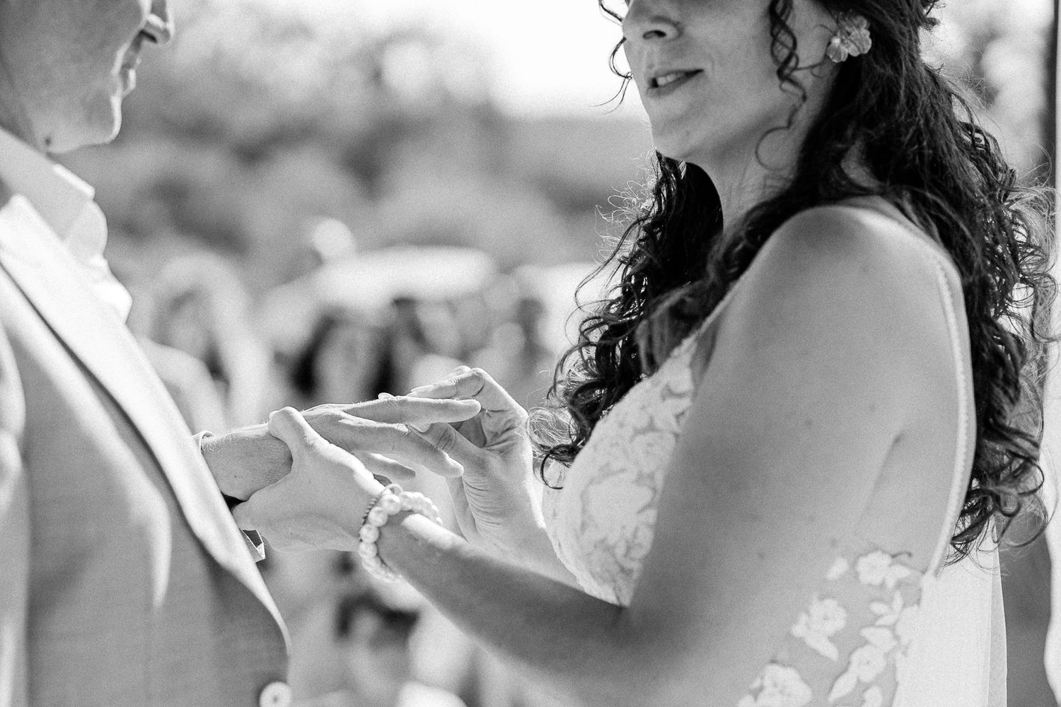 Black and white photo of bride placing wedding ring on partner's finger during outdoor ceremony.