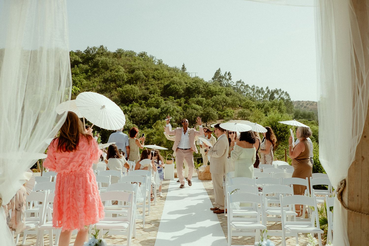 Outdoor wedding ceremony with guests holding white parasols walking down an aisle lined with white chairs.