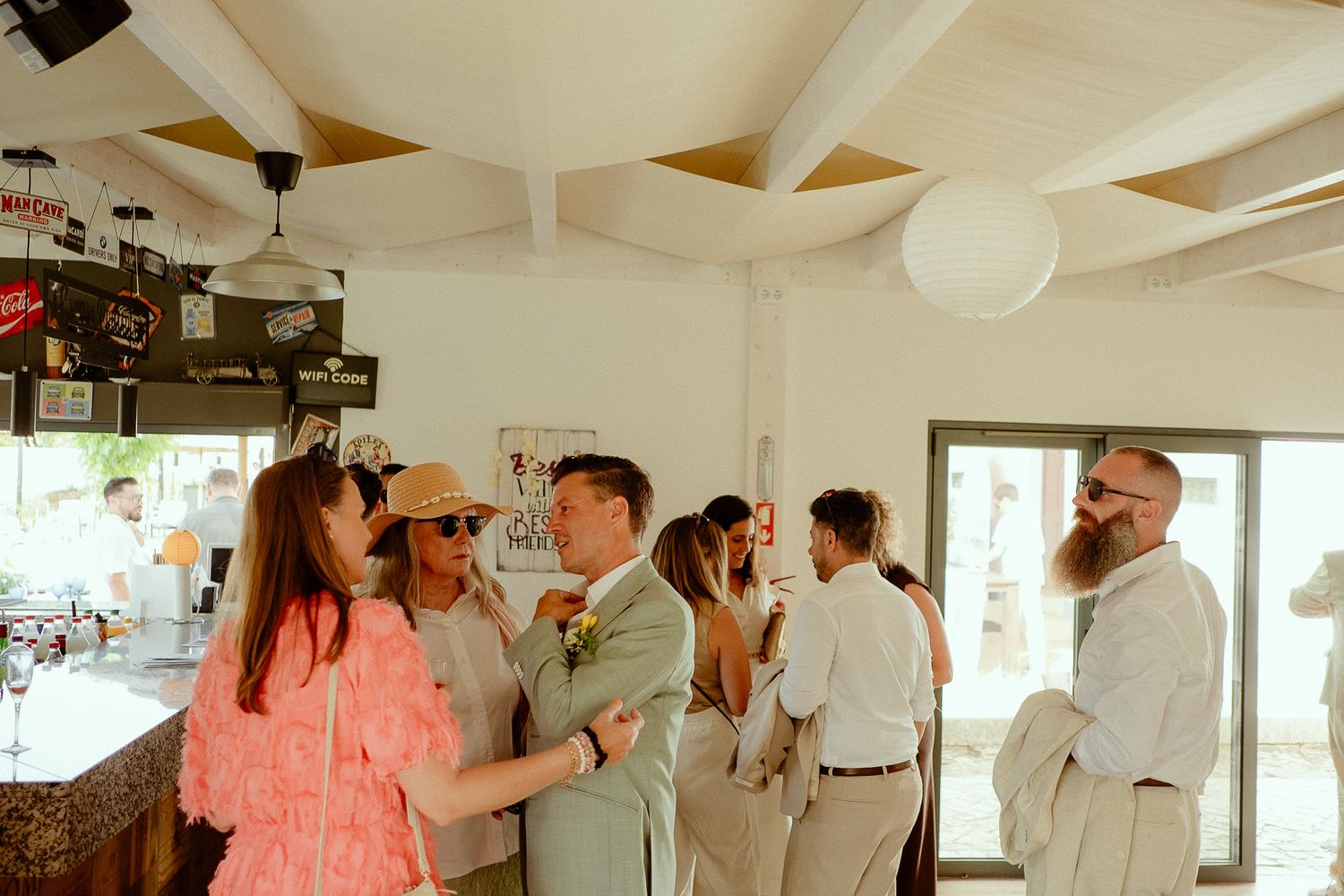 Guests mingle at an outdoor wedding reception in a bright venue, with the groom in a sage suit greeting attendees.