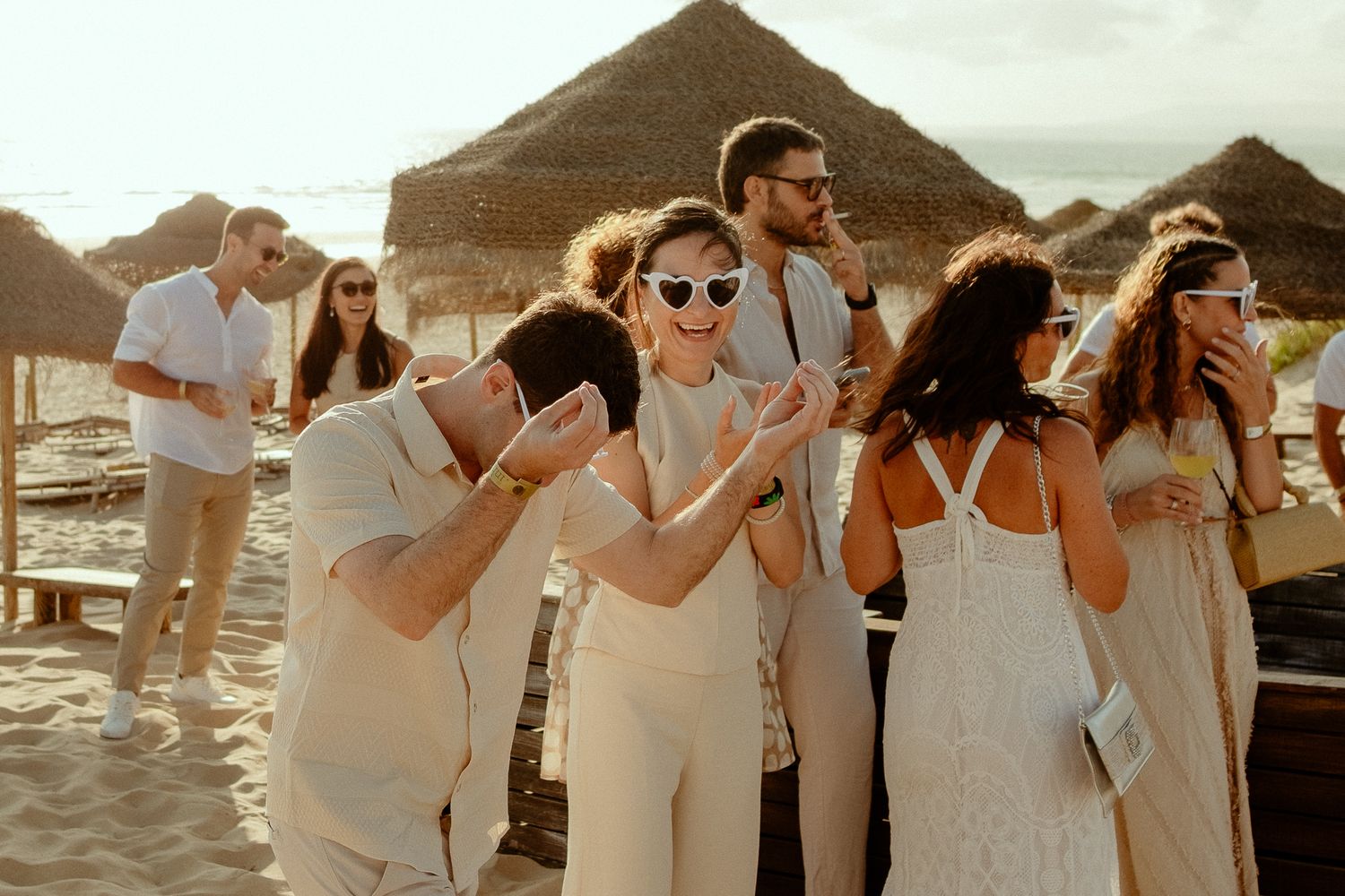 Guests in white outfits celebrate at a beach wedding reception with thatched umbrellas in the background.
