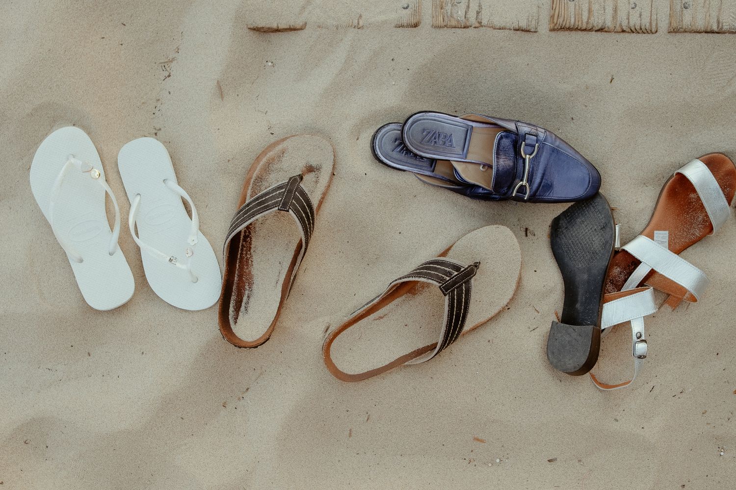 Four pairs of sandals and flip-flops arranged on sandy beach, including white, brown, blue, and tan styles.