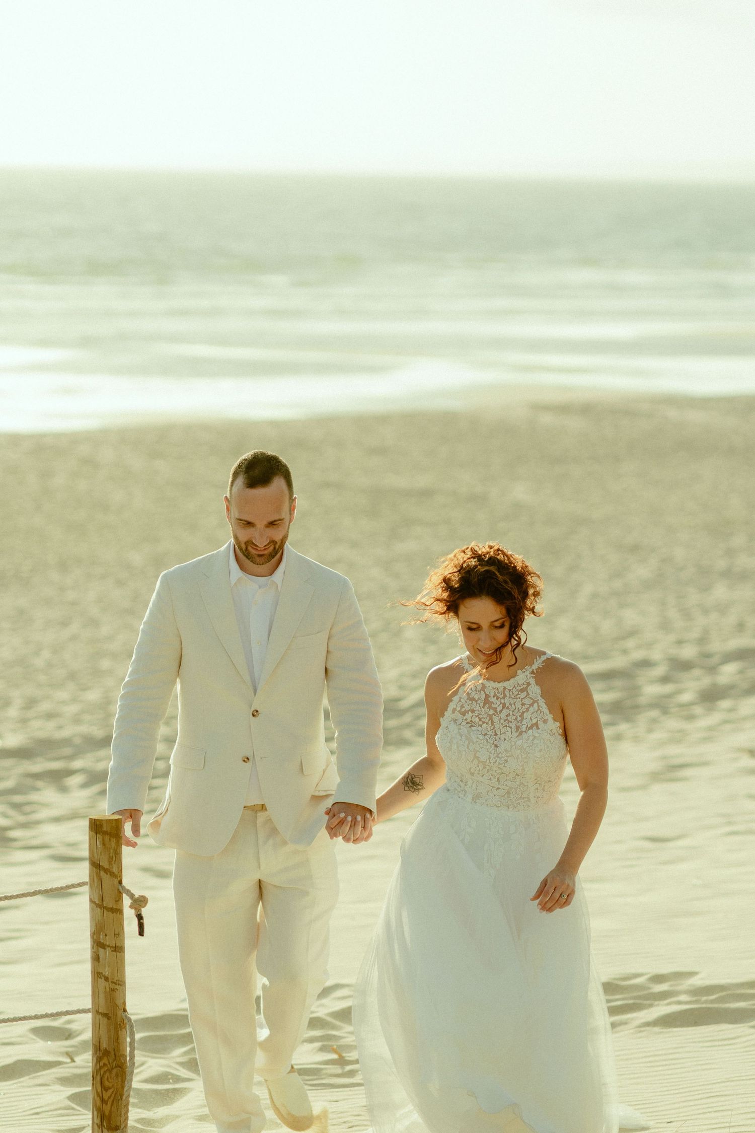Bride and groom walking hand in hand on a sandy beach at golden hour, wearing white wedding attire.