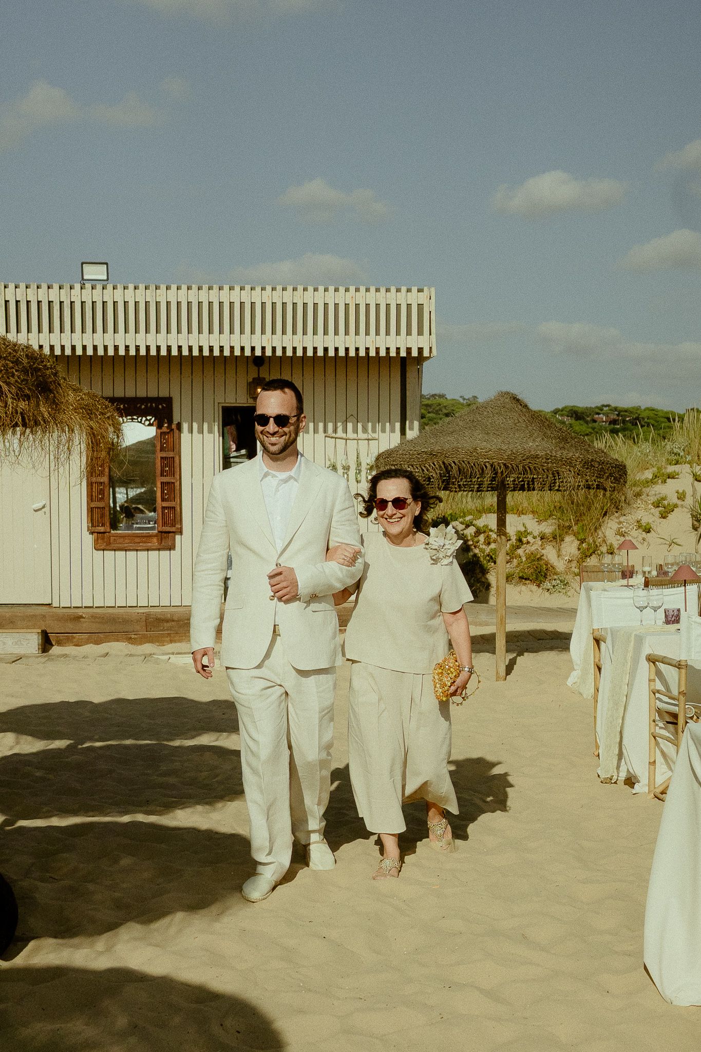 Groom in white linen suit walks with older woman in beige outfit on sandy beach wedding venue.