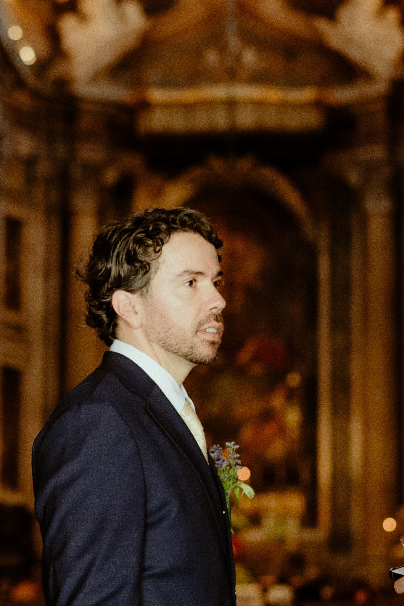 A man in a dark suit with a boutonniere stands in profile inside an ornate church during a wedding ceremony.