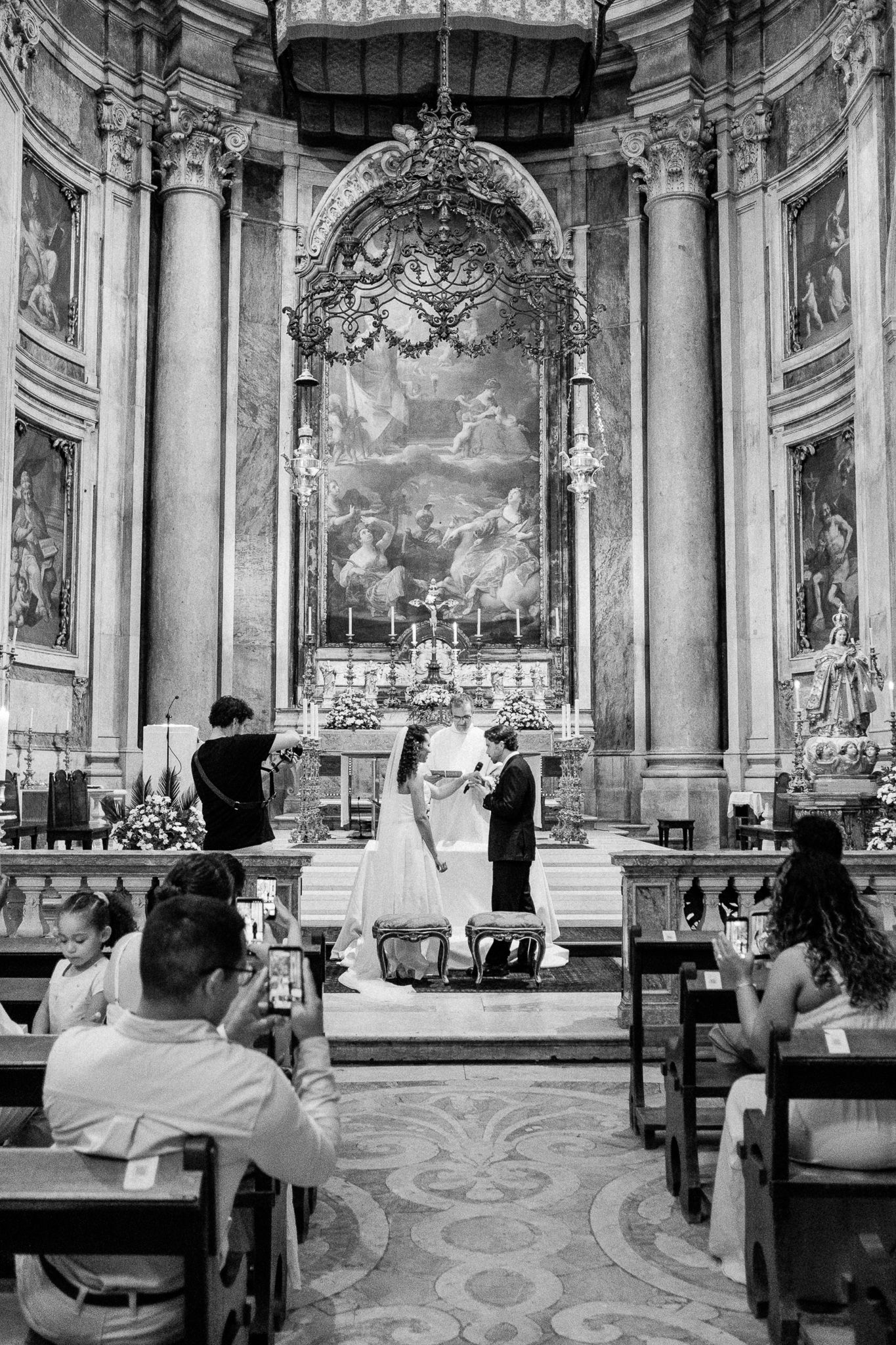 Black and white photo of a wedding ceremony inside an ornate Italian baroque church with grand altar artwork.