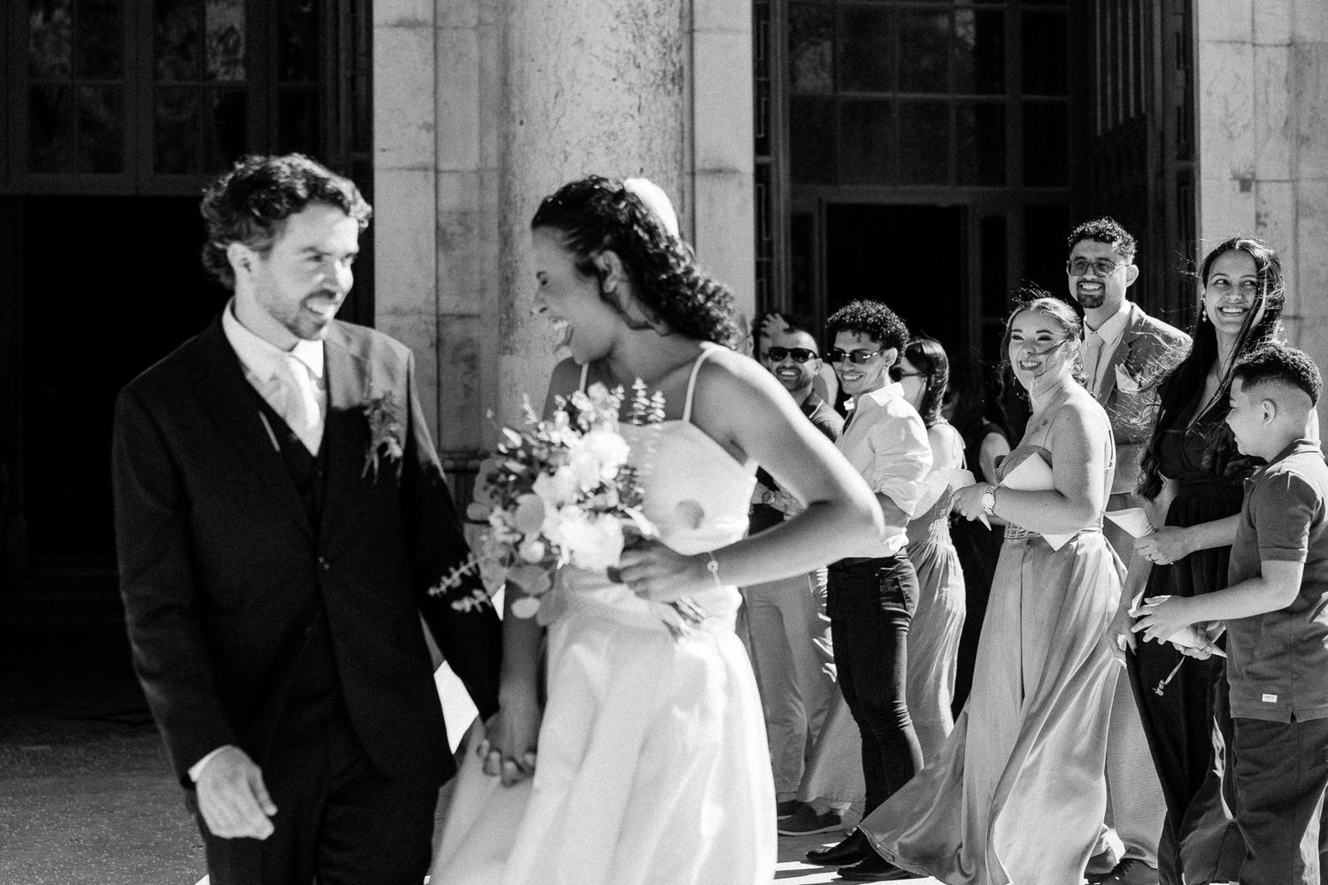 Black and white wedding photo of bride and groom smiling, surrounded by joyful guests outside a grand building.