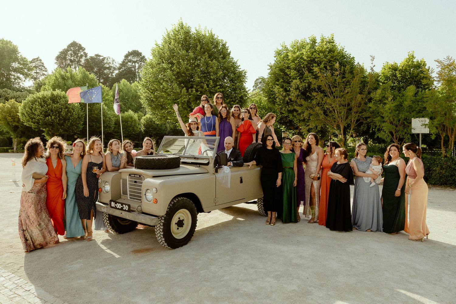 Large wedding party posing with a vintage Land Rover outdoors, surrounded by lush green trees.