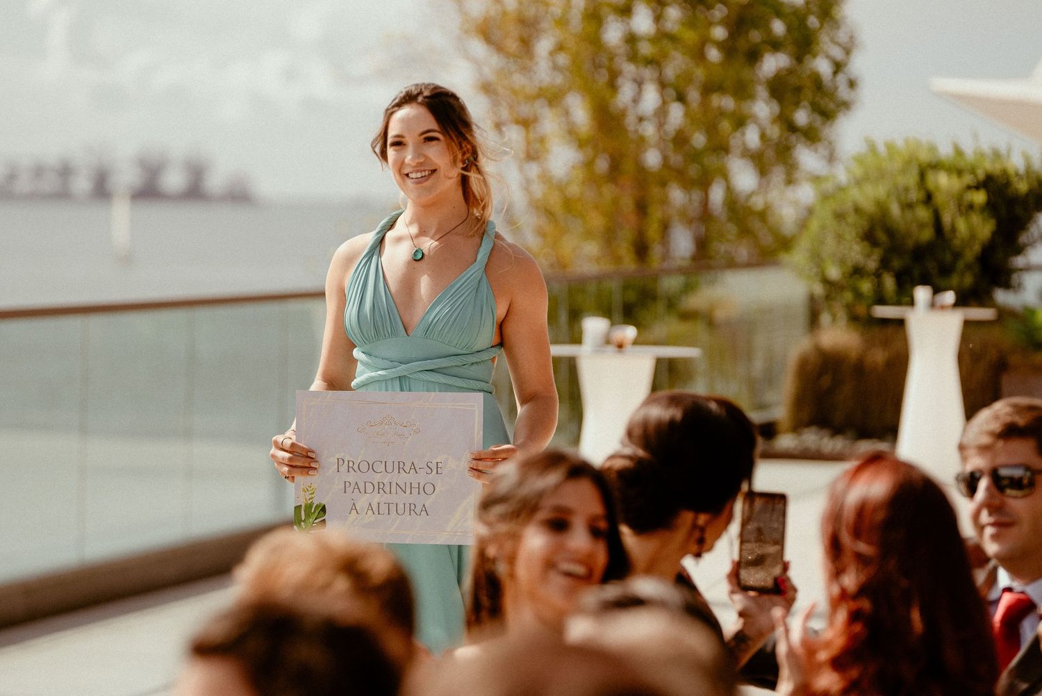 A happy woman in a blue dress holds a coffee cup while engaging with children seated at classroom desks.
