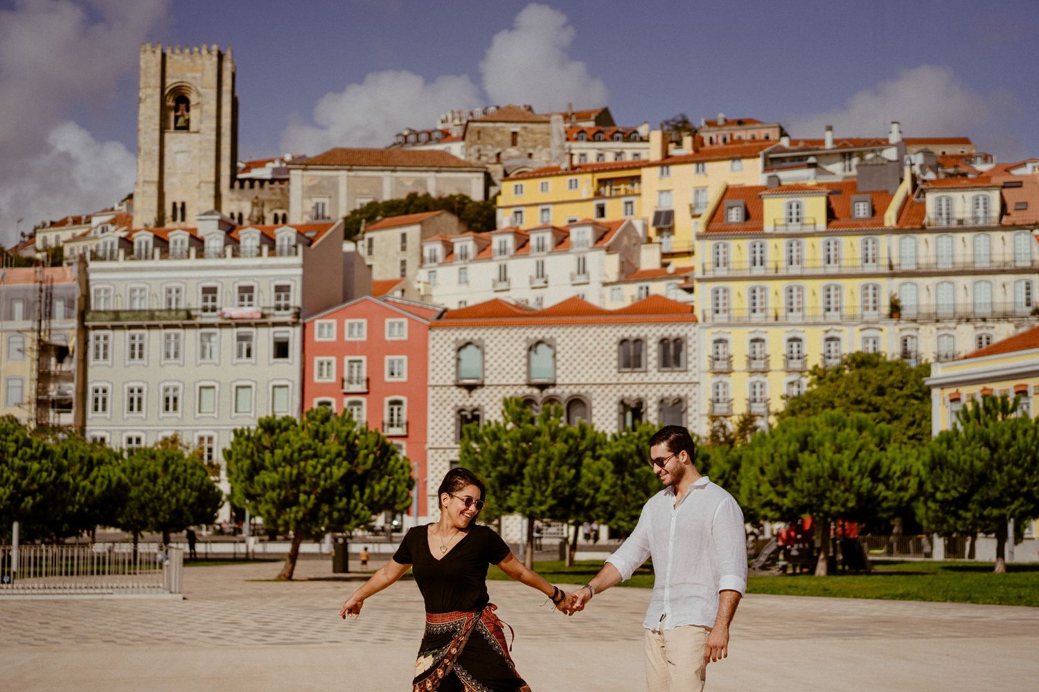 People walk through Lisbon's colorful Alfama district with pastel buildings and historic architecture in the background.