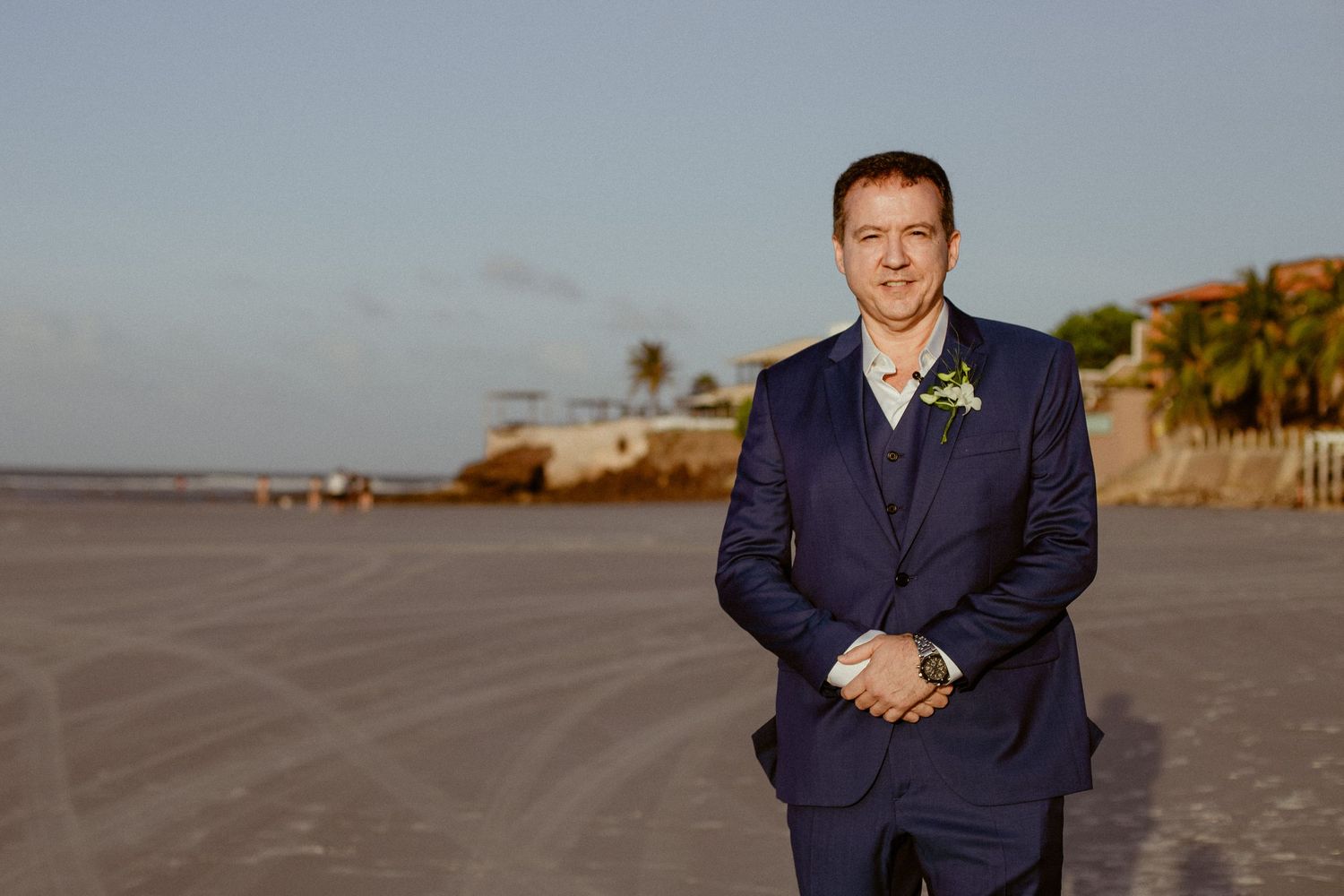A person wearing a navy blue suit stands on an empty beach at sunset with waves in the background.