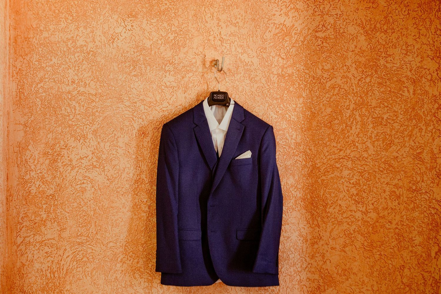 A navy blue blazer with satin lapels and a light-colored boutonniere hangs against an orange textured background.
