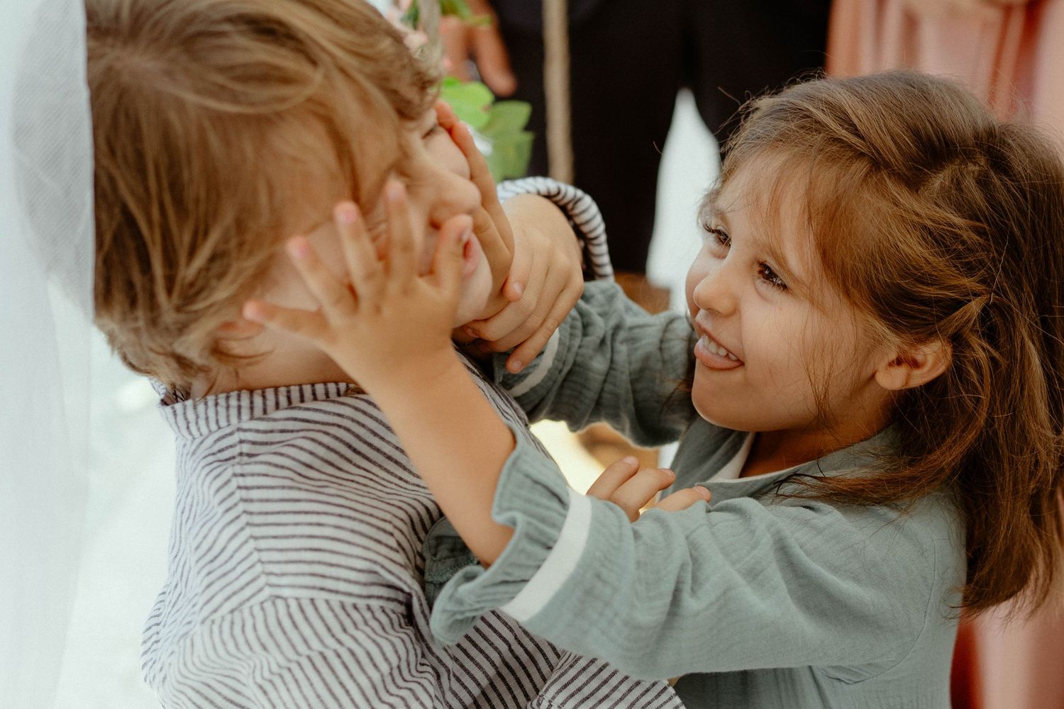 Two children playing and laughing together while one squishes the other's cheeks playfully.