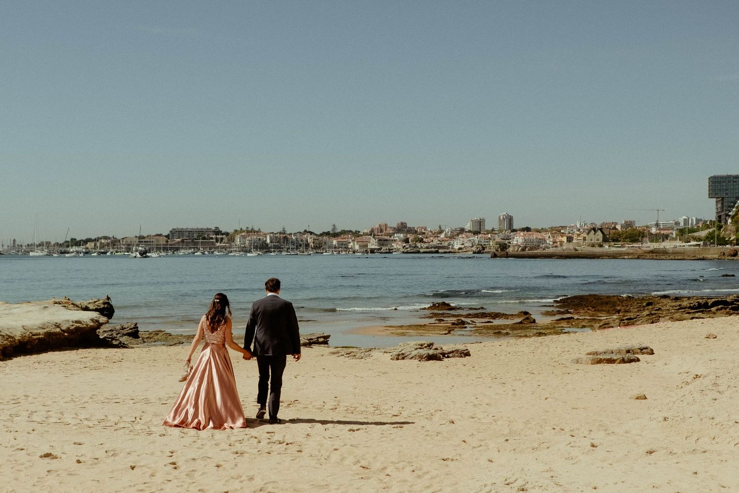 Two people walk along a scenic beach with driftwood and coastal town views in the background.