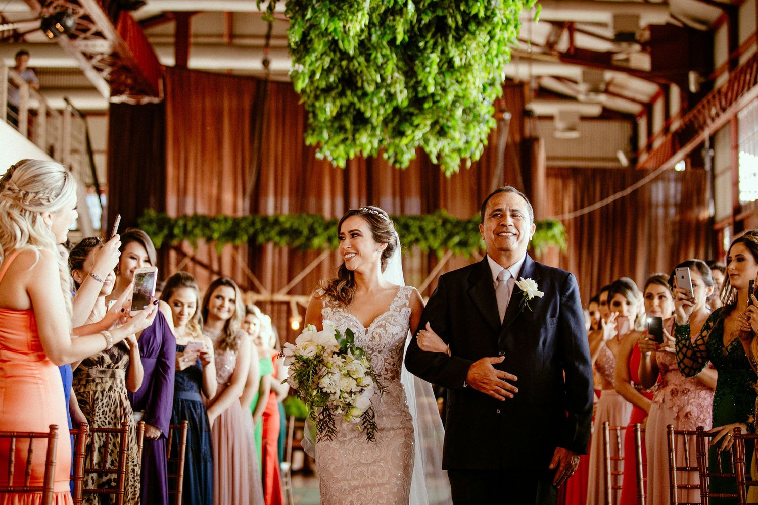 Wedding ceremony processional in venue decorated with hanging greenery and warm lighting.