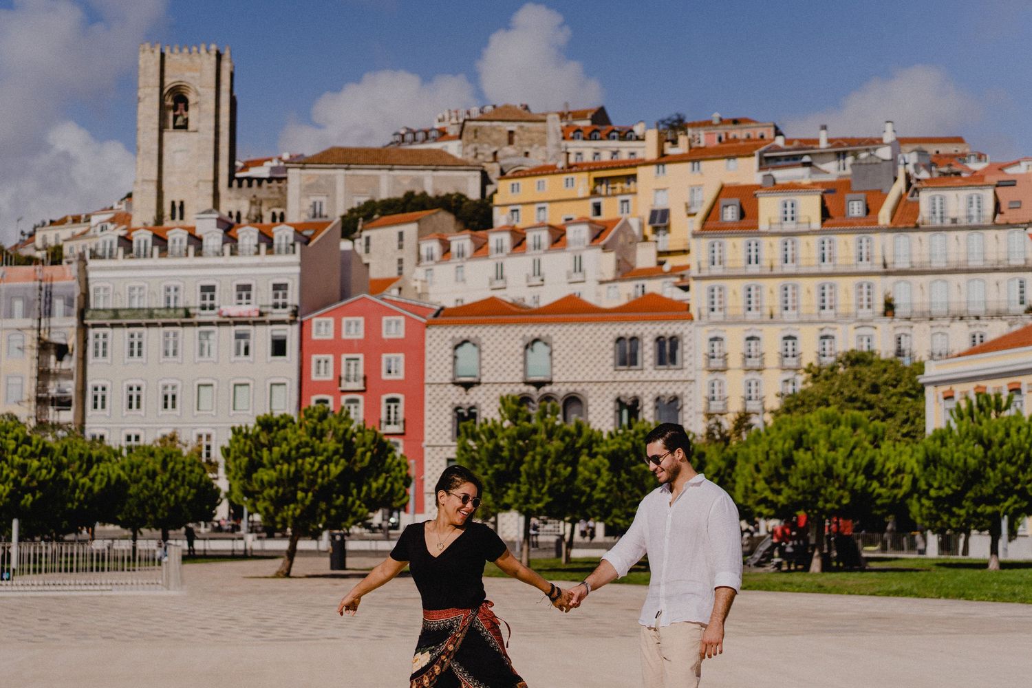 Couple dancing in front of colorful historic buildings and a tower in Lisbon's city center on a sunny day.