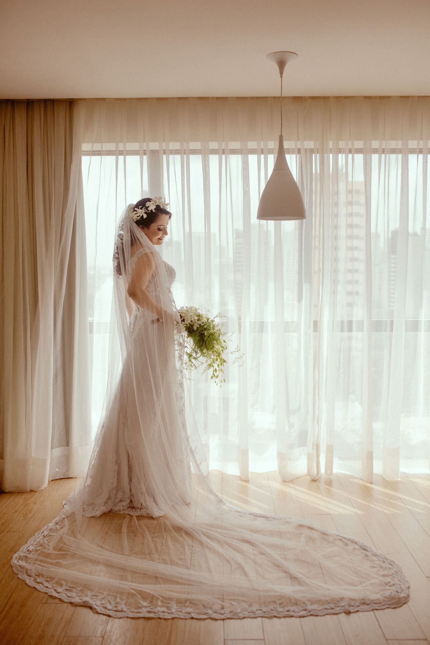 A bride in an elegant white lace wedding gown with long train stands by sheer curtains holding a delicate bouquet.