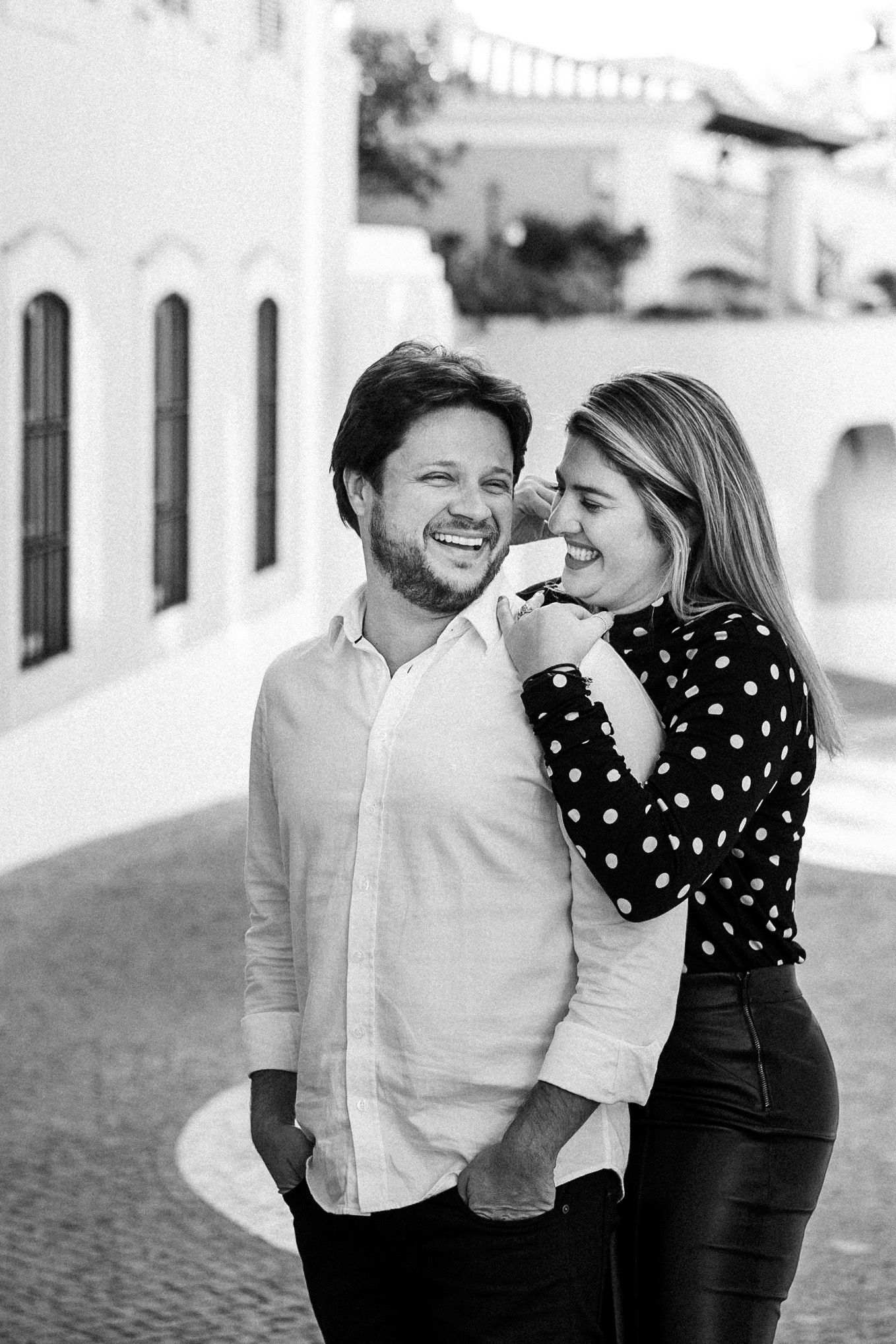 A romantic black and white photo of a couple sharing a joyful laugh together on a street with white buildings.
