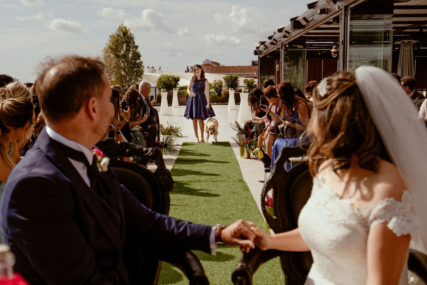A scenic outdoor wedding ceremony takes place on a grassy lawn with guests gathered on both sides of the aisle.