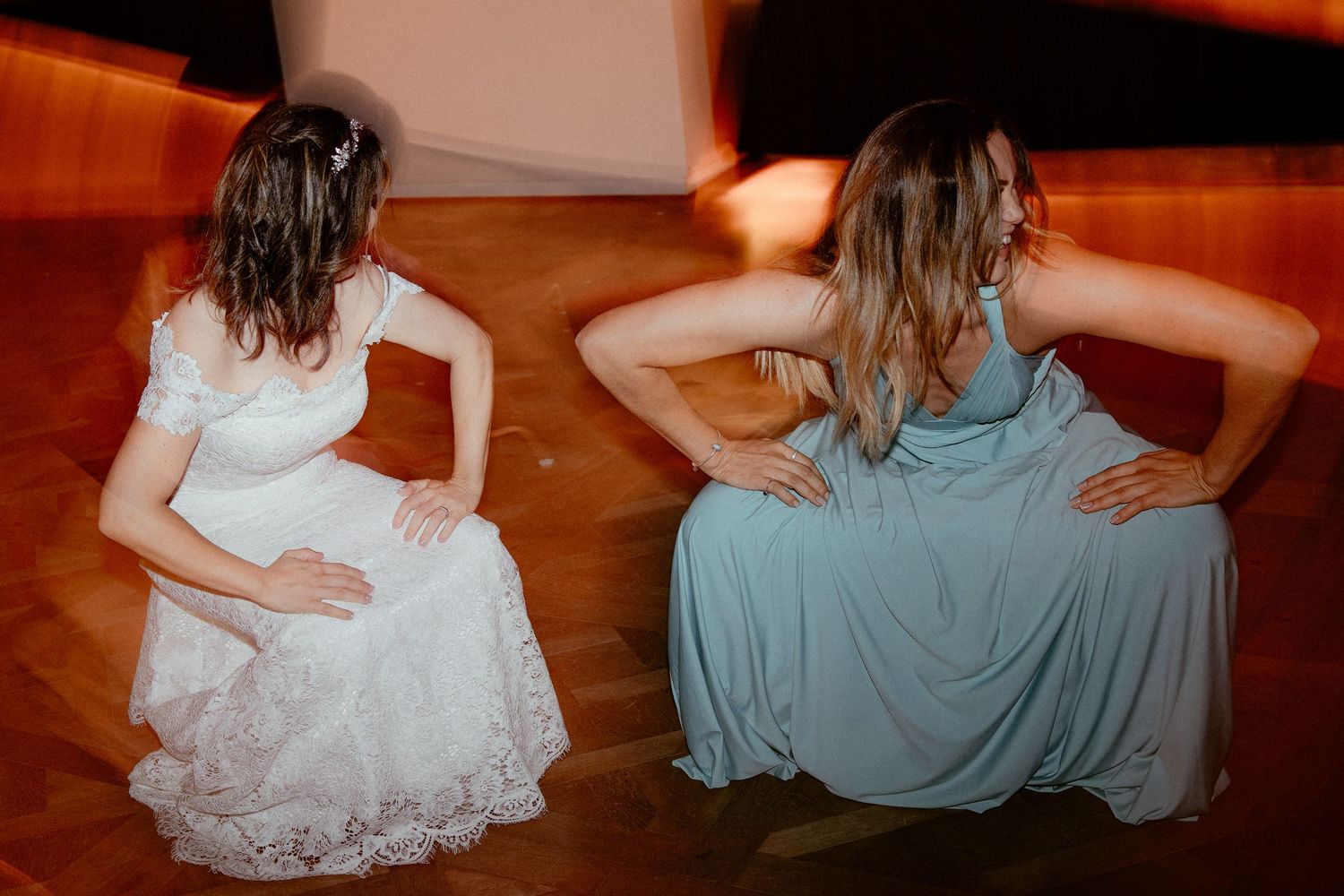 Two people in elegant dresses sit back-to-back on polished wooden floor creating symmetrical poses.