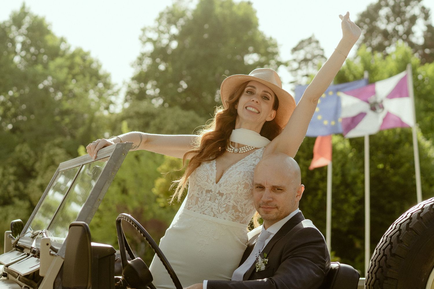 Joyful bride in white dress and hat raises arm while sitting with groom in vintage convertible car outdoors.
