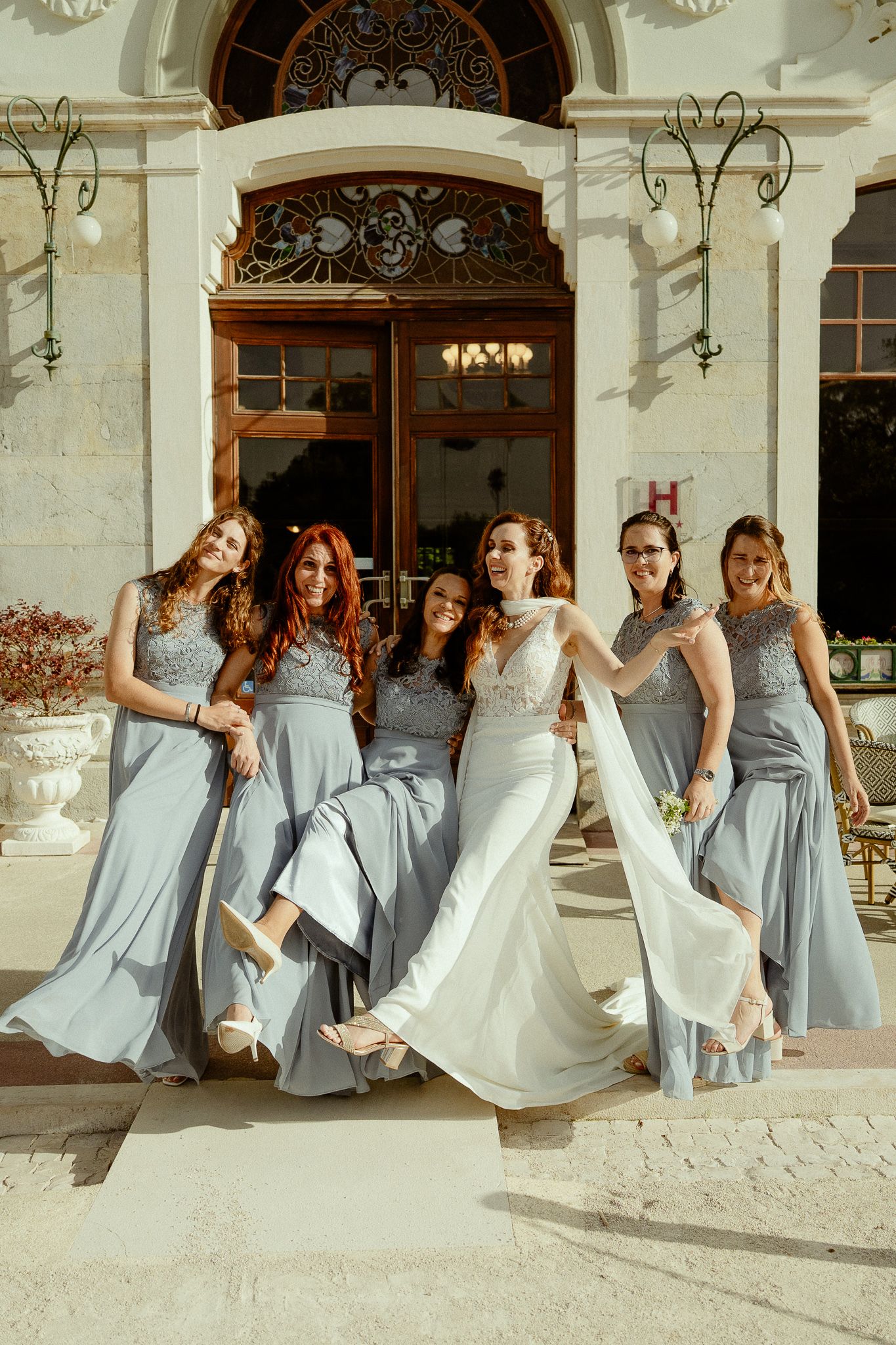 Bride in white gown poses with five bridesmaids in matching gray dresses outside elegant venue.