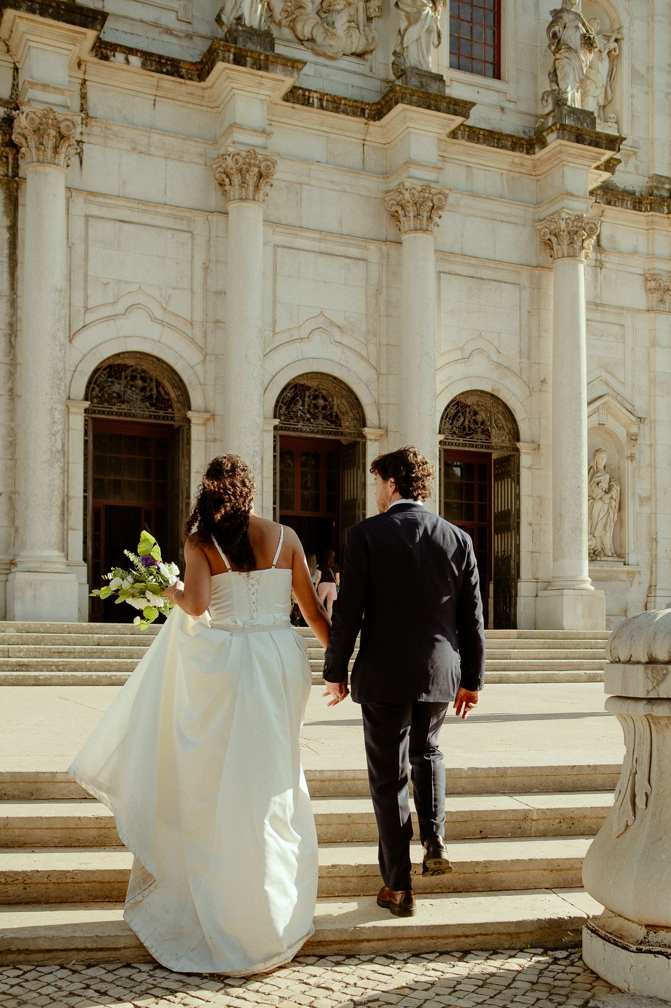 Bride and groom walking up stone steps toward ornate white baroque church entrance on their wedding day.