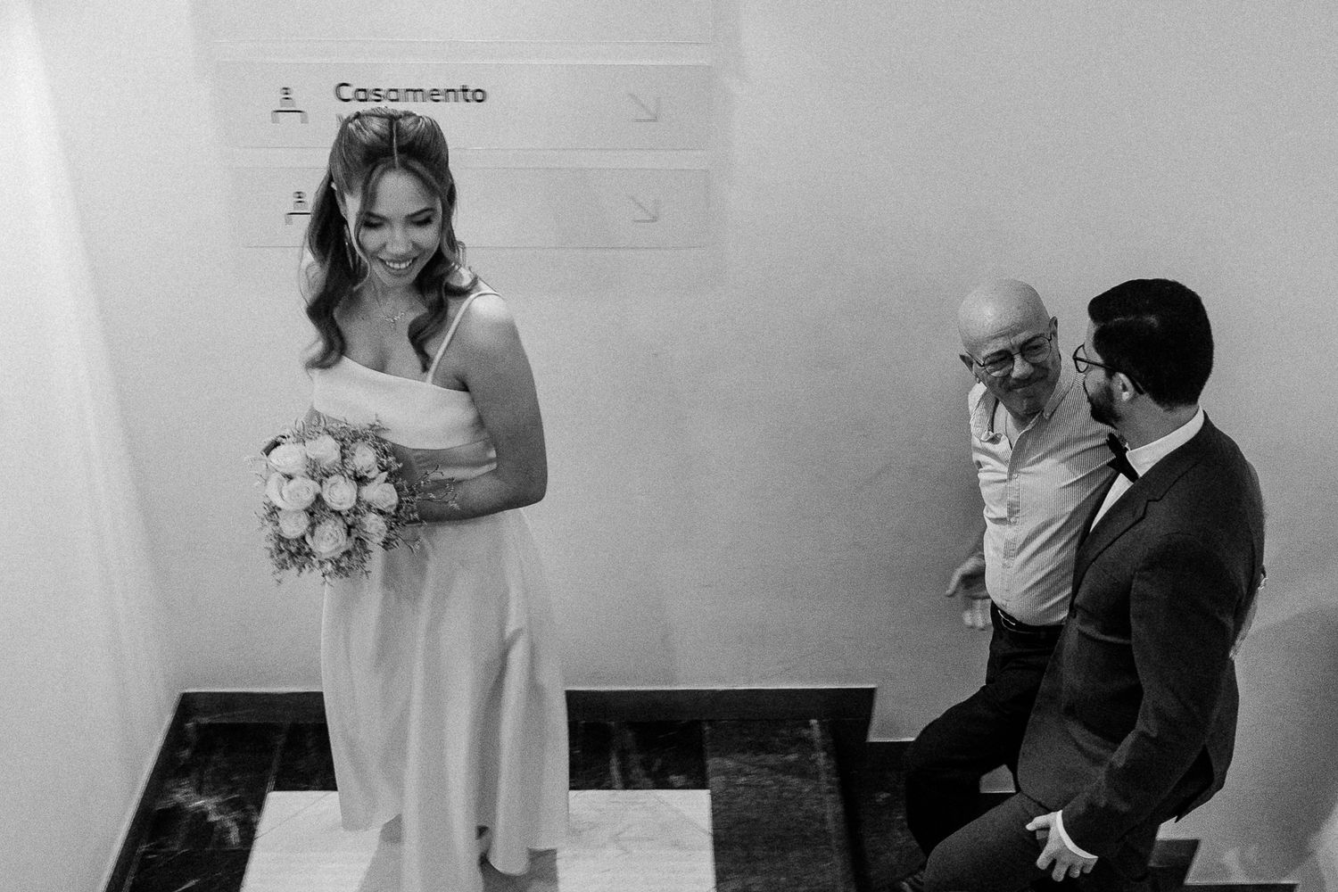Black and white photo of a smiling bride holding a bouquet at a wedding ceremony with two men nearby.