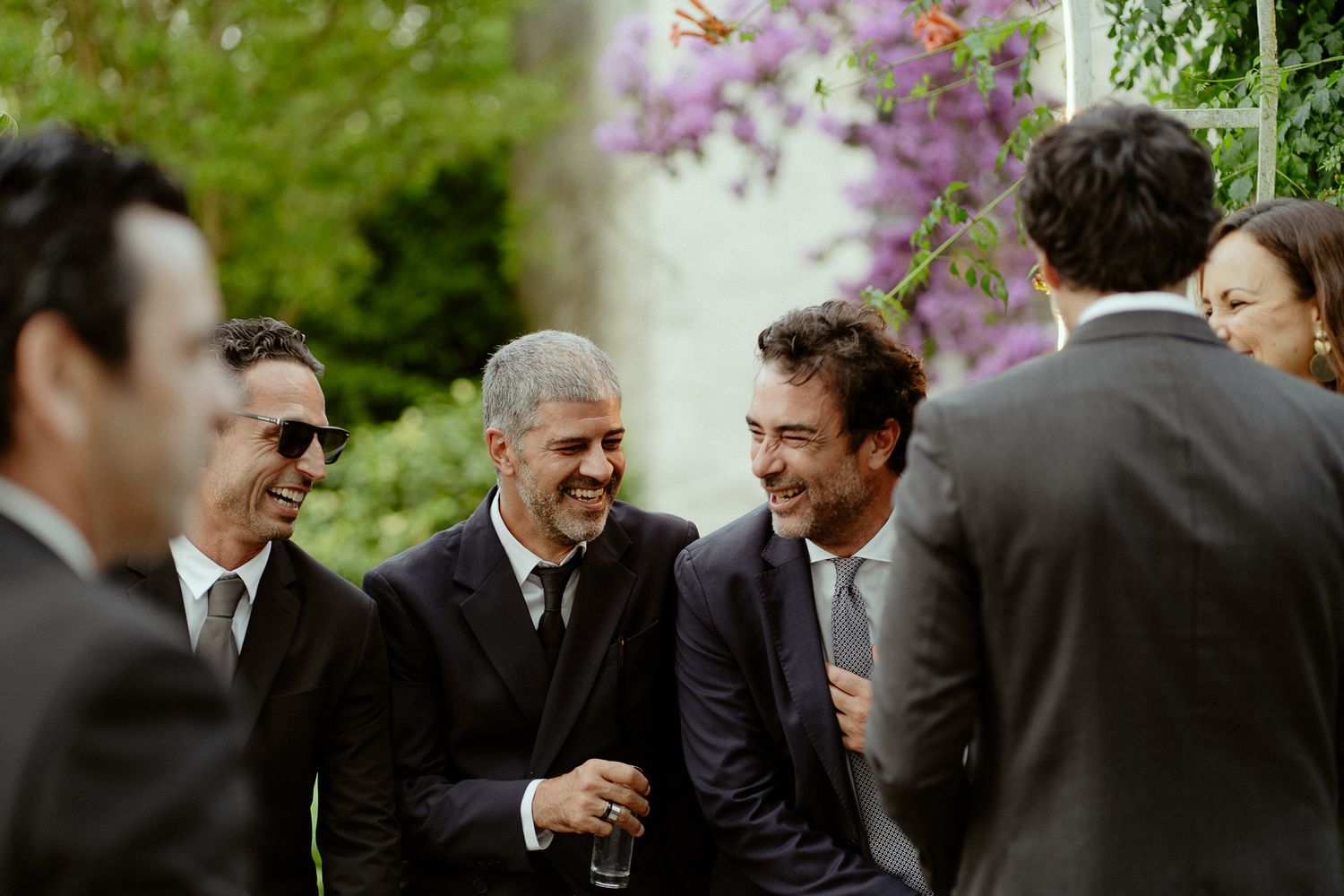 Group of men in dark suits laughing and socializing outdoors at a formal event with purple flowers in background.